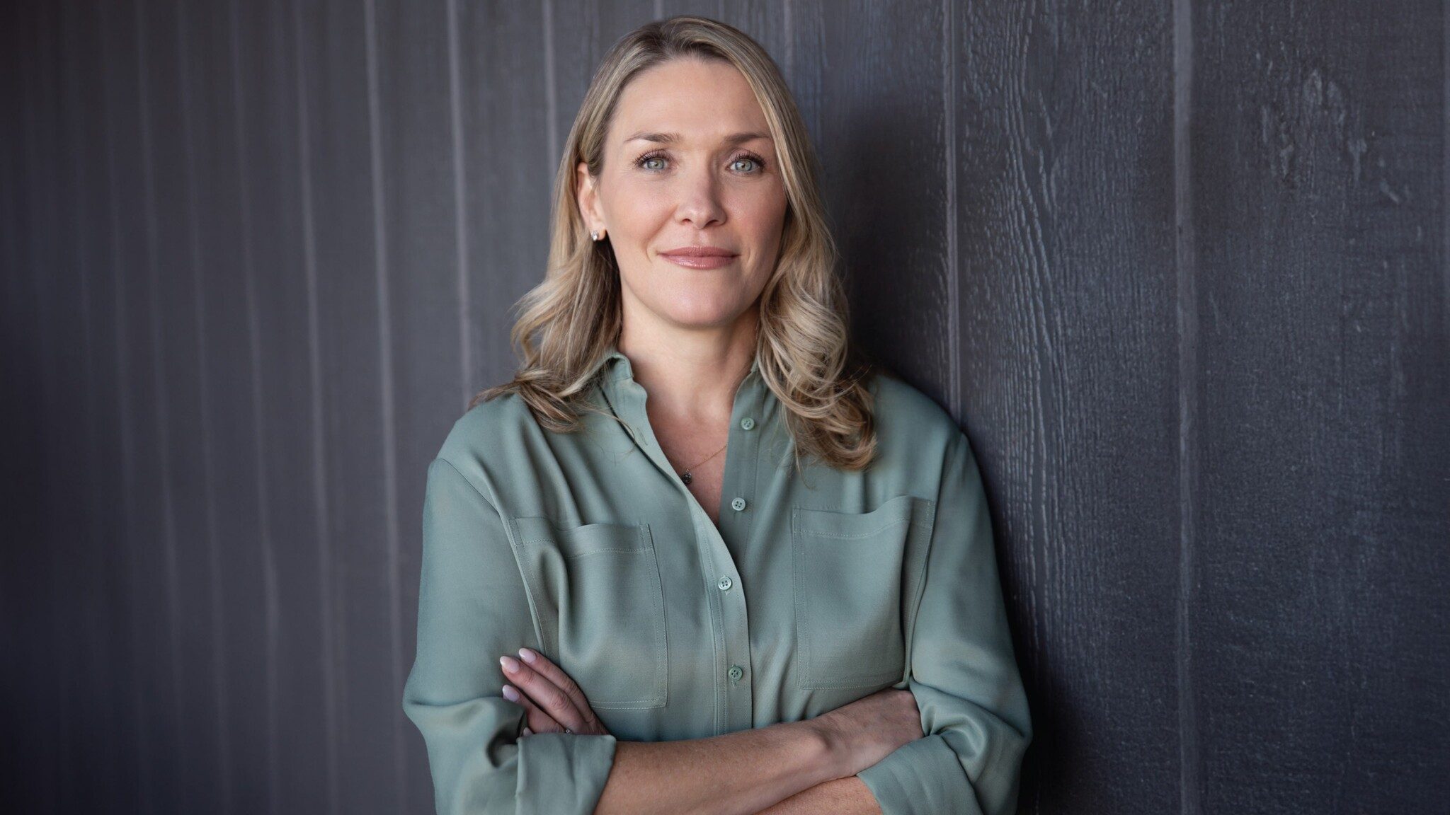 A professional woman with blonde hair wearing a green shirt stands crossed arms, smiling slightly, against a dark gray background.