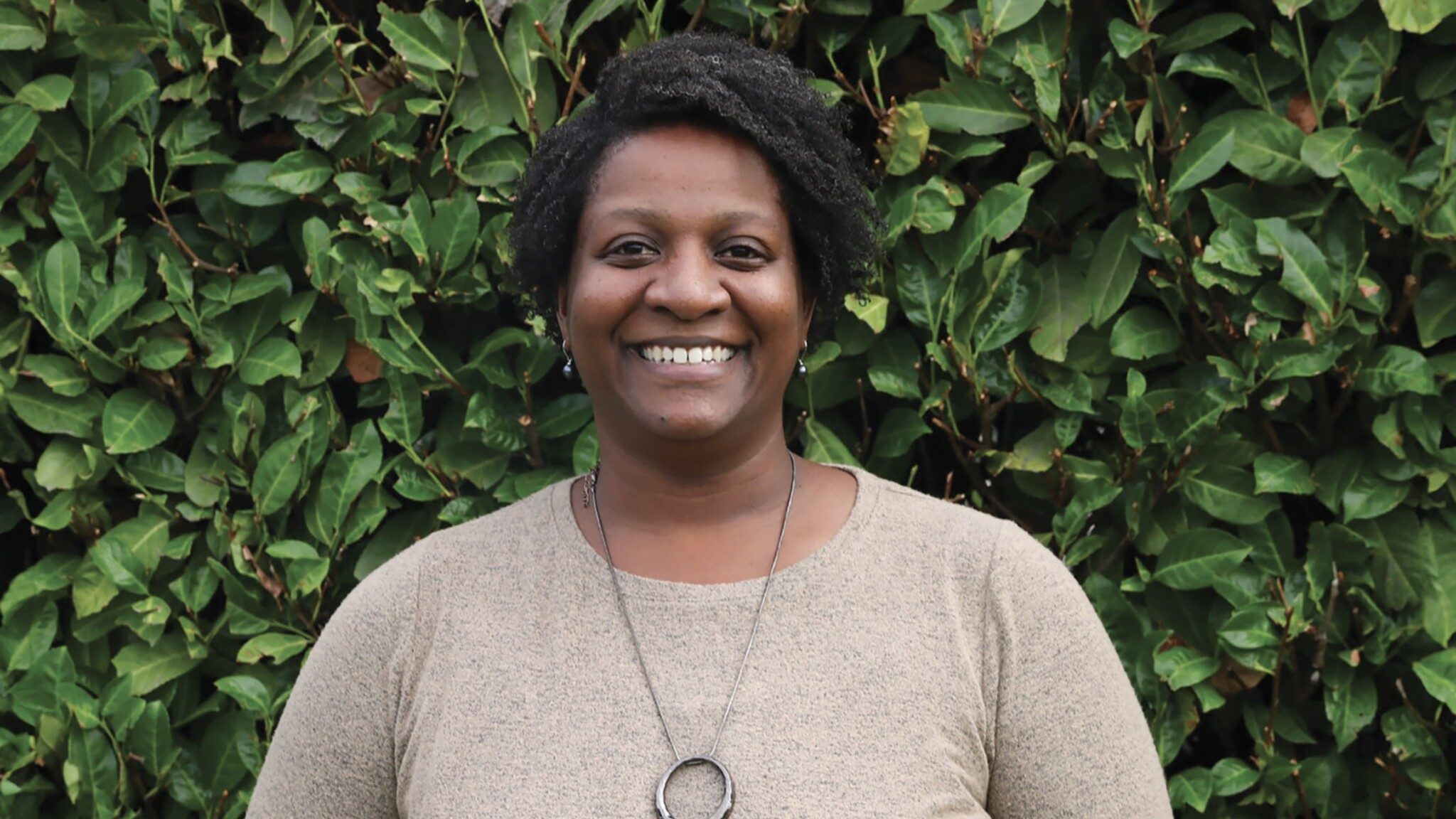 A smiling woman with short curly hair standing in front of a lush green ivy-covered wall. she is wearing a beige sweater and a necklace.