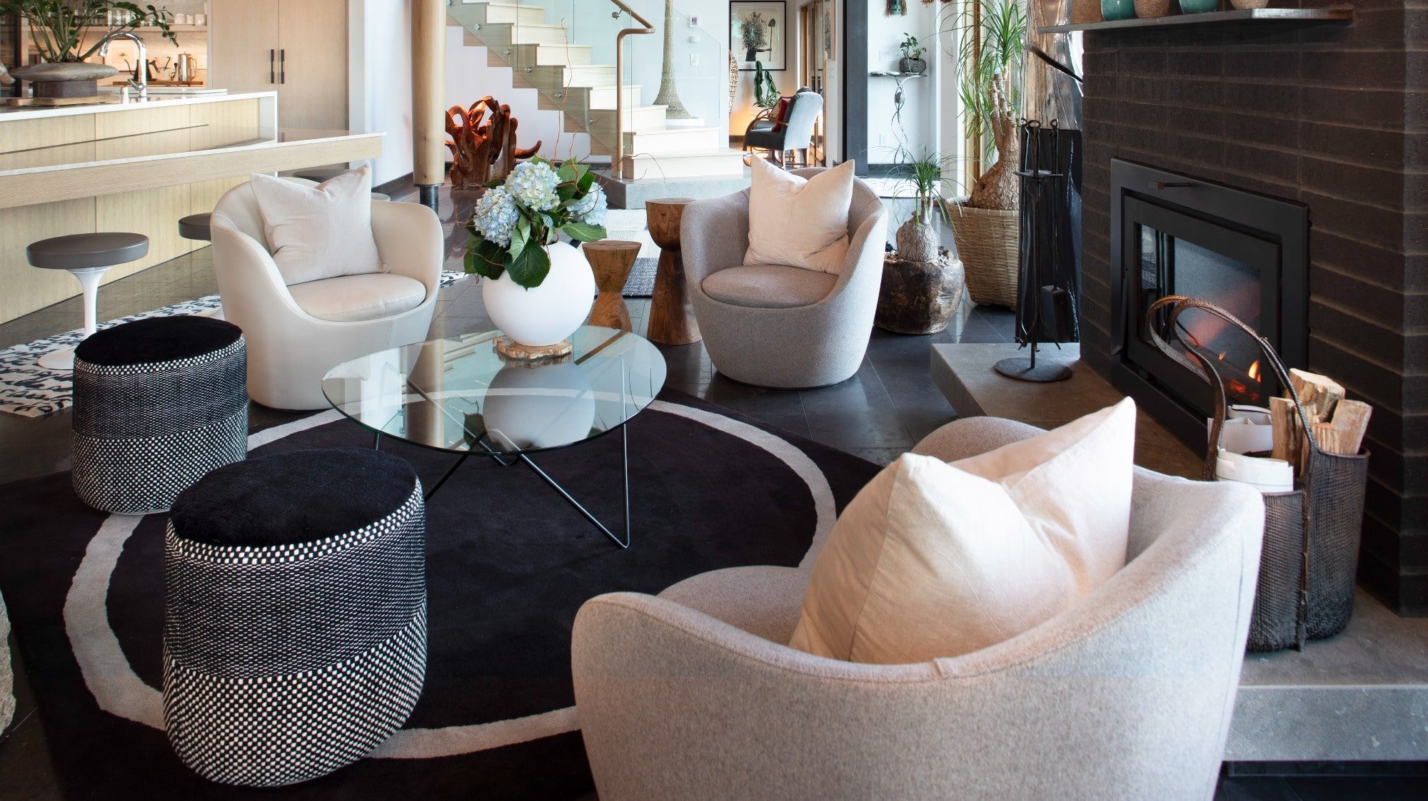 Modern living room with stylish furniture, including plush chairs, a glass coffee table, and a fireplace, featuring elegant decor and an open kitchen in the background.