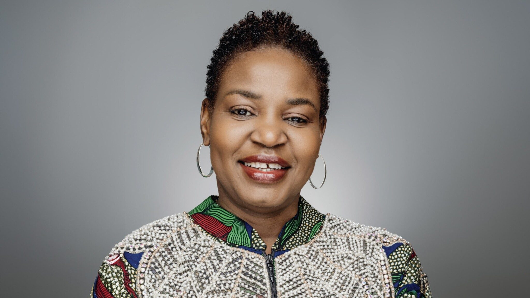 Portrait of a smiling black woman with short hair, wearing a colorful jacket and hoop earrings against a gray background.