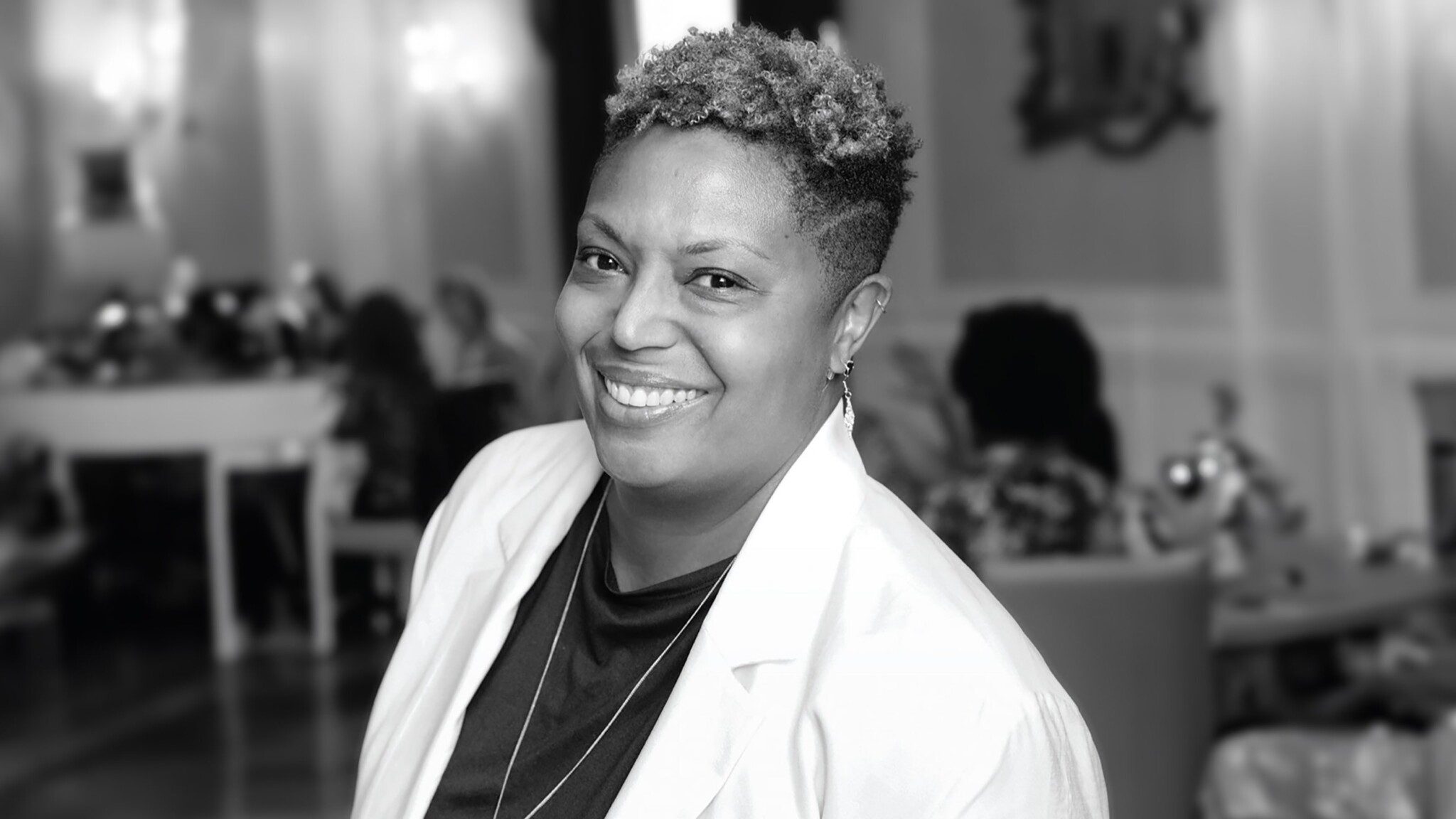 A black woman with short curly hair, smiling at the camera in a black and white photograph, wearing a white jacket over a dark top, with a blurred indoor background.