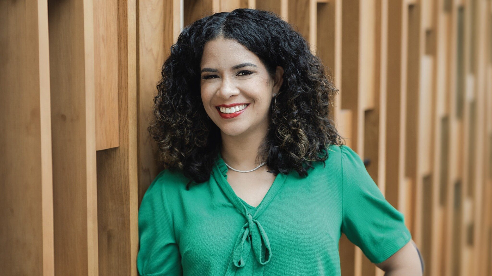 A woman with curly hair, wearing a green blouse, smiles in front of a wooden slatted background.