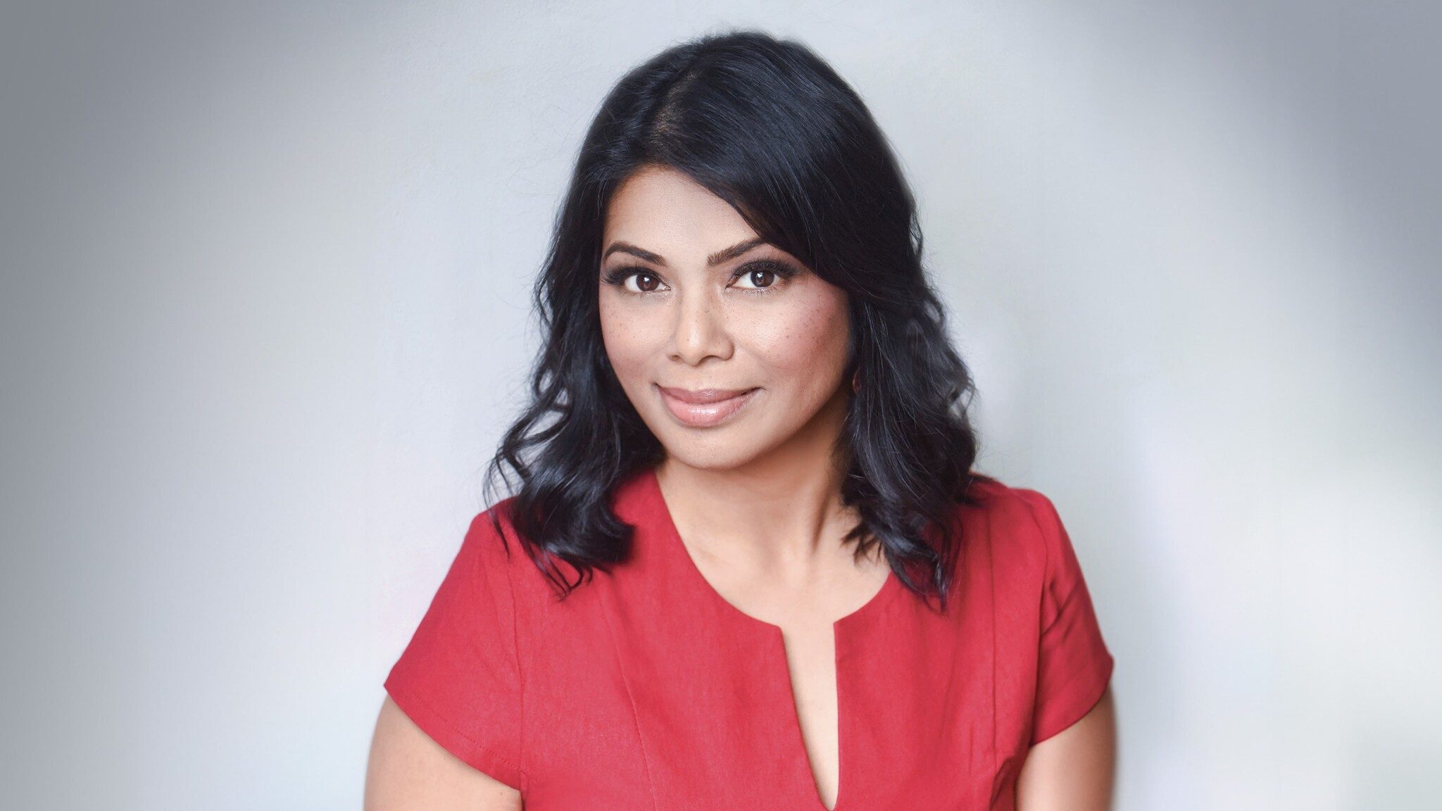 Portrait of a woman with dark hair and a confident smile, wearing a red blouse against a soft, neutral background.