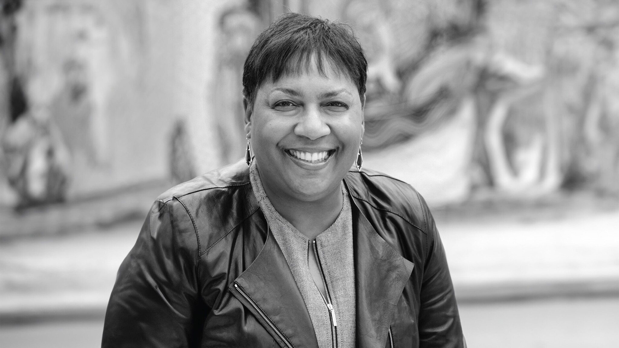 Black-and-white portrait of a smiling woman in a leather jacket, sitting outdoors with a blurred urban background.