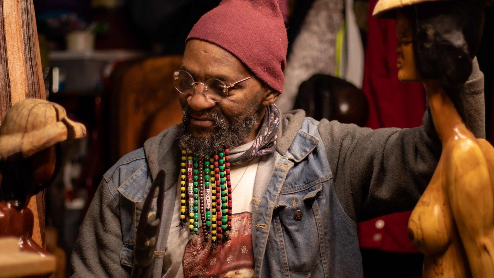 A bearded man in glasses and a red beanie smiles with ingenuity while working in a cluttered workshop surrounded by wooden sculptures.