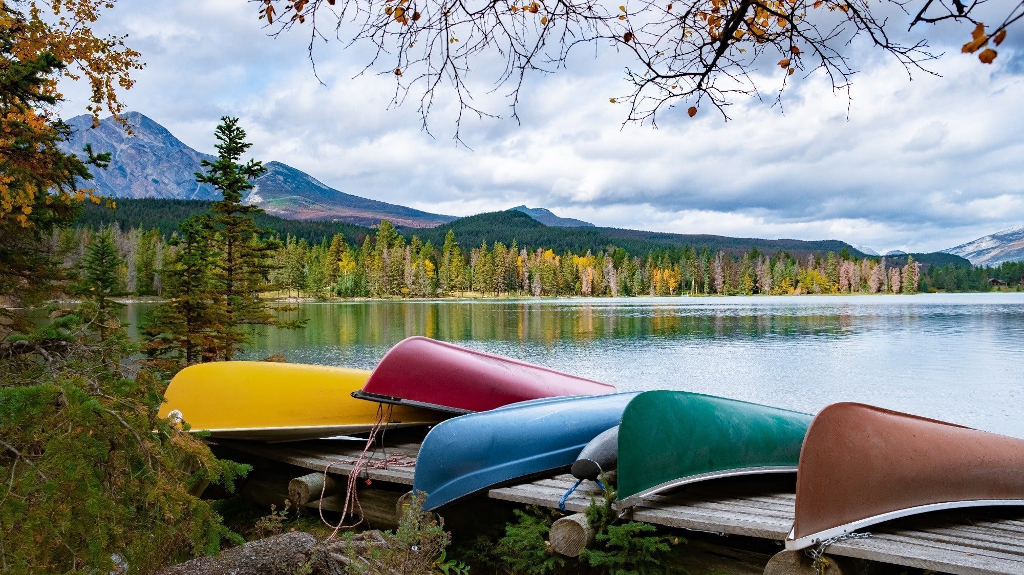 Colorful canoes stacked on a wooden dock beside a serene lake, with autumn trees and mountains in the background, creating an atmosphere of peace.