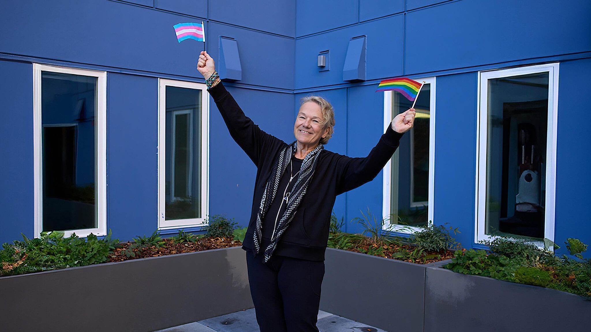 A person stands outside a blue Senior Center, holding small transgender pride and LGBTQ+ pride flags, smiling with arms raised as the center celebrates the LGBTQ+ community.