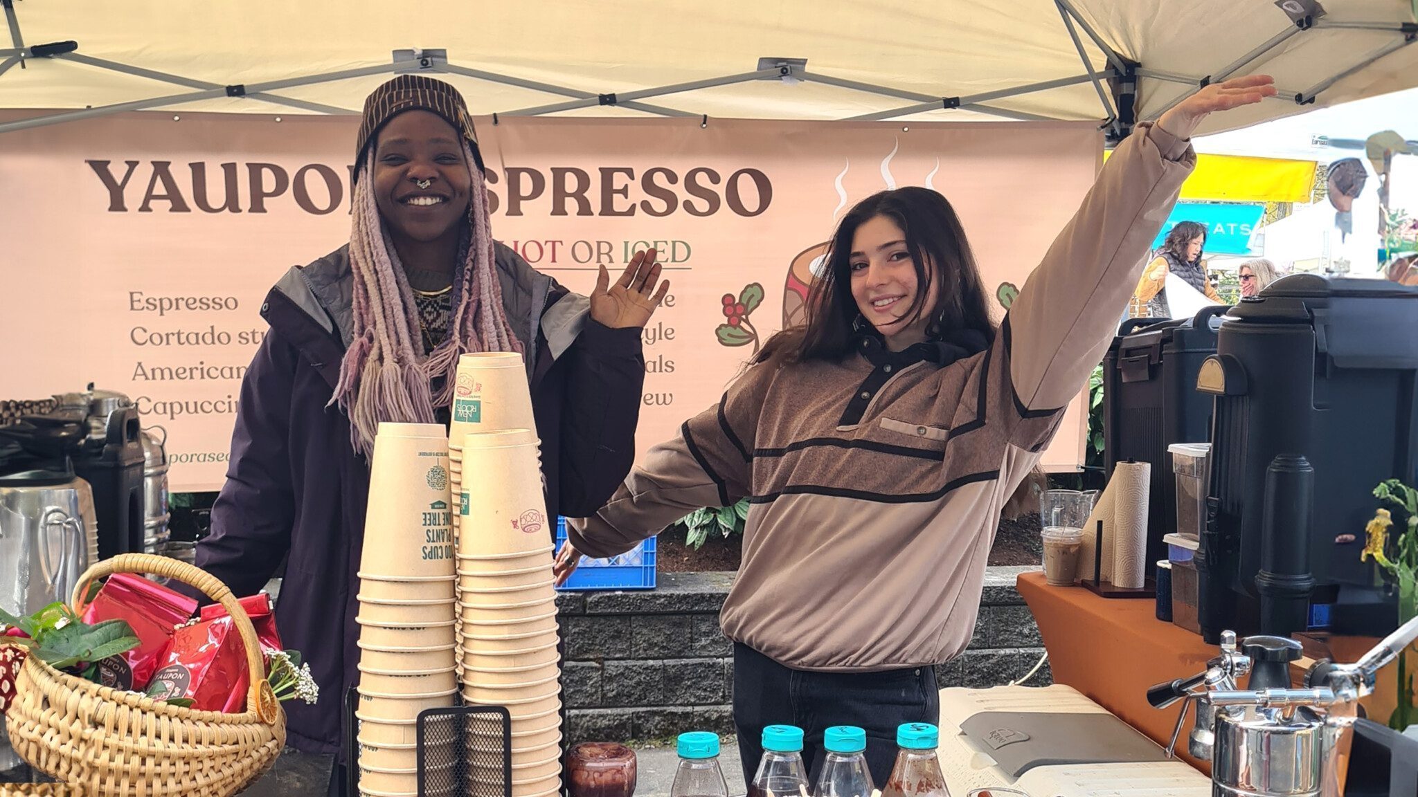 Two people stand smiling under a tent at a coffee stand. They are surrounded by coffee cups, bottles, and a basket with flowers. A banner behind them reads "Yaupon Espresso Presents." The diverse diaspora of flavors includes their specialty, the American Americano.