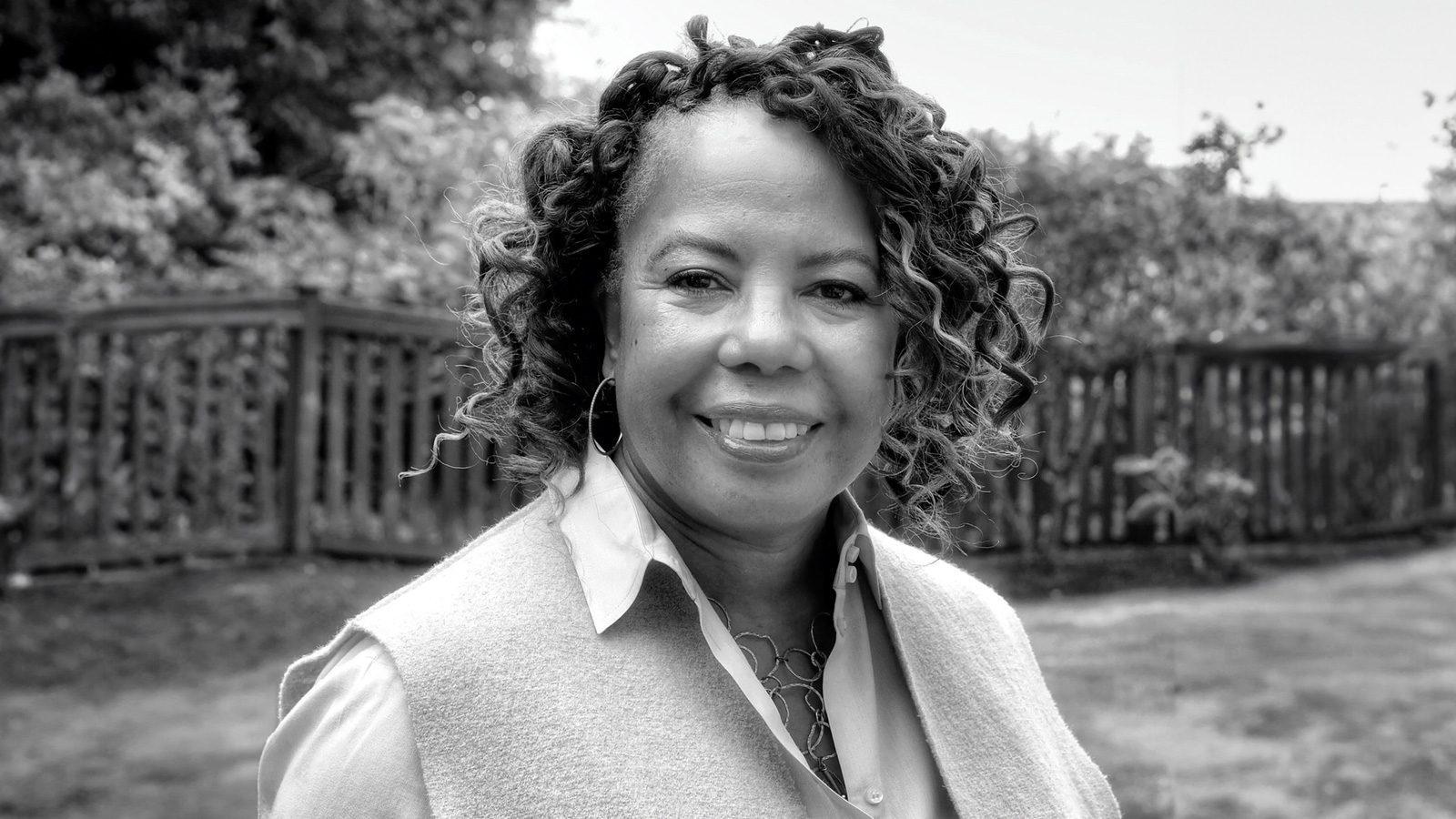 A trailblazing woman with curly hair smiles while standing outside in front of a wooden fence, captured beautifully in black and white.