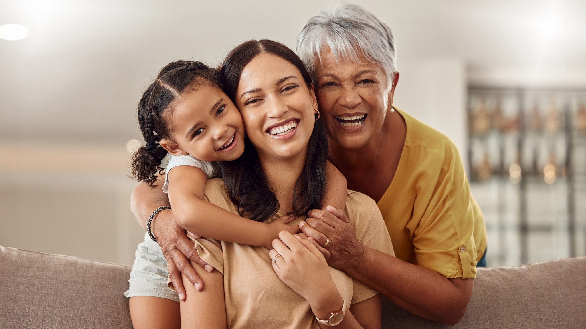 A smiling child hugs a young woman, who sits in front of an older woman, on a couch. All three are close together, with the older woman resting her hands on the young woman's shoulders, embodying the unspoken burden of caregiving shared across generations.
