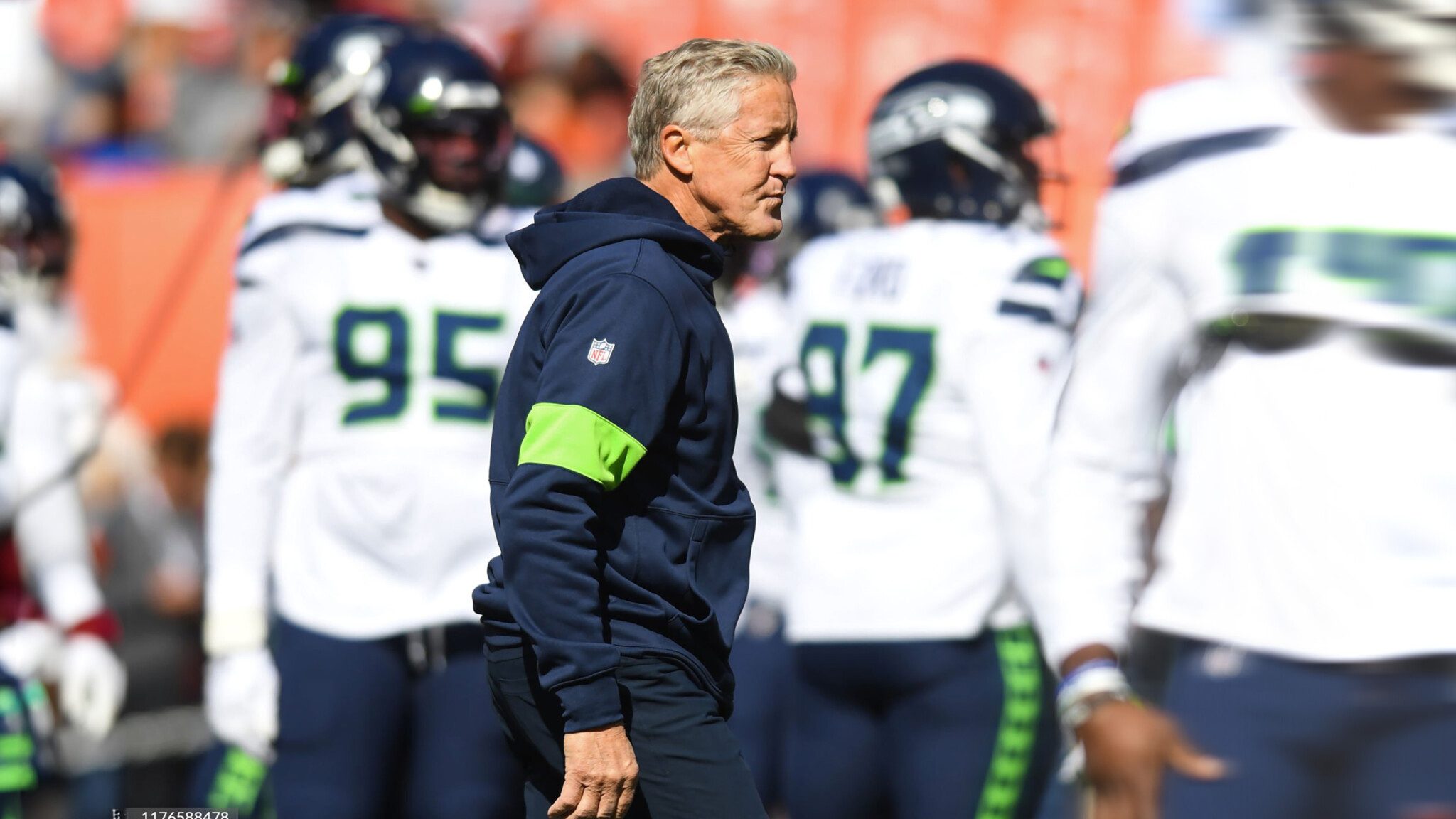 A coach in a navy hoodie and green armband, reminiscent of Pete Carroll's quirky legacy, walks on the football field while players in white and blue uniforms stand in the background.