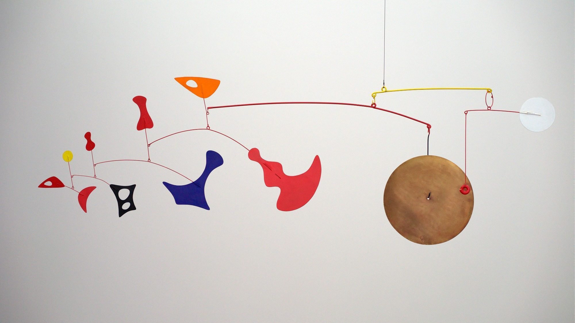 A colorful hanging mobile with abstract shapes, including red, yellow, orange, blue, and white pieces, balances delicately connected to thin red and yellow rods against a white background. The dynamic design is reminiscent of the Call of Calder’s engaging artistry.