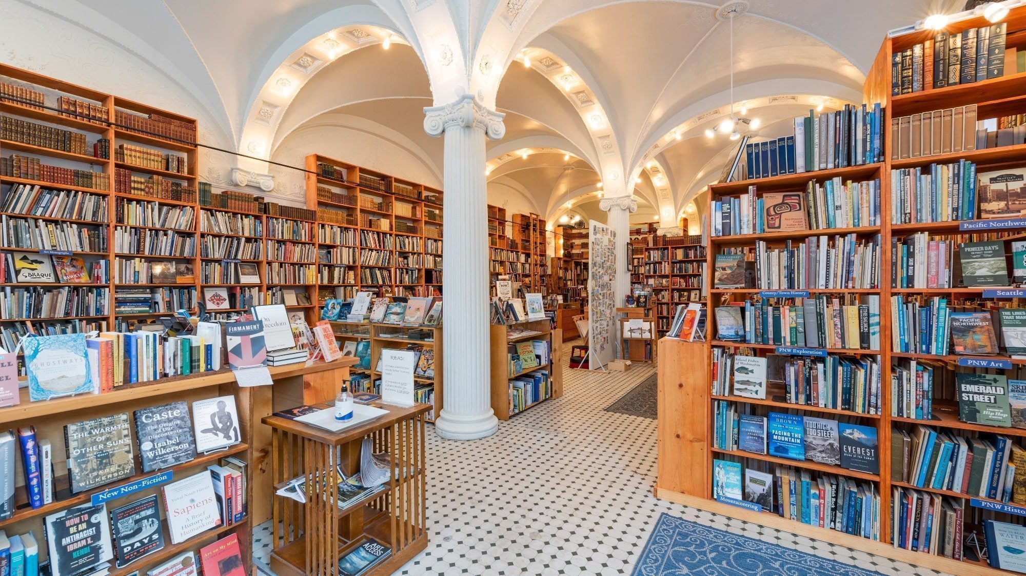 A spacious bookstore with tall bookshelves filled with various books. Sunshine highs stream through large windows, enhancing the ambiance. An arched ceiling with ornate details adds to the charm, while a pillar stands in the center of the store.