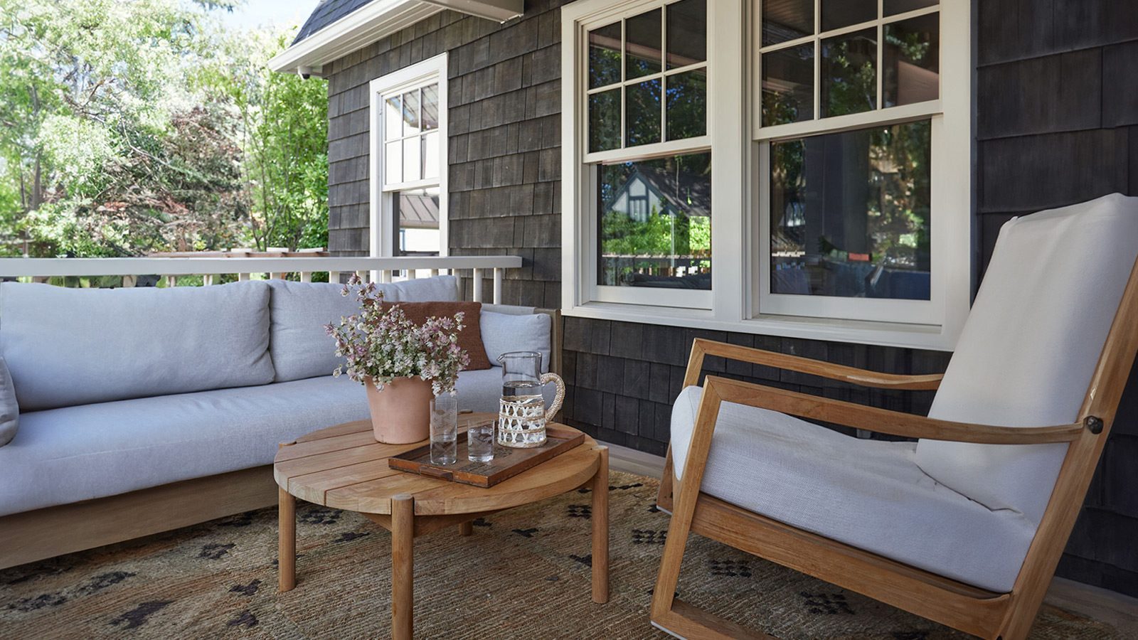 An outdoor patio for maximalists features a cushioned sofa, a wooden rocking chair, and a round wooden coffee table with a potted plant and glassware, all under the serene Montlake sky.