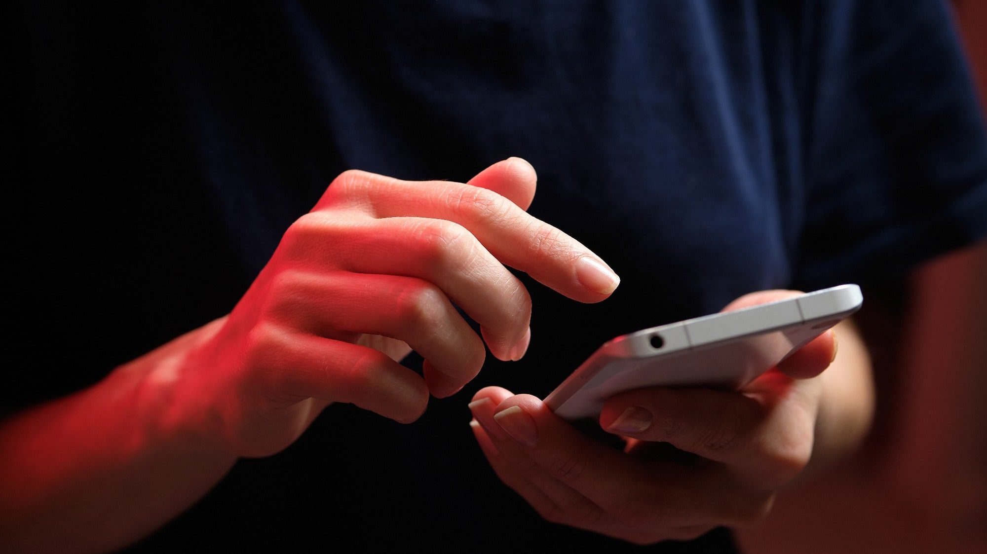 A person in a dark shirt is holding and using a smartphone with both hands, illuminated by red lighting, showcasing the bad habit of always checking your phone.