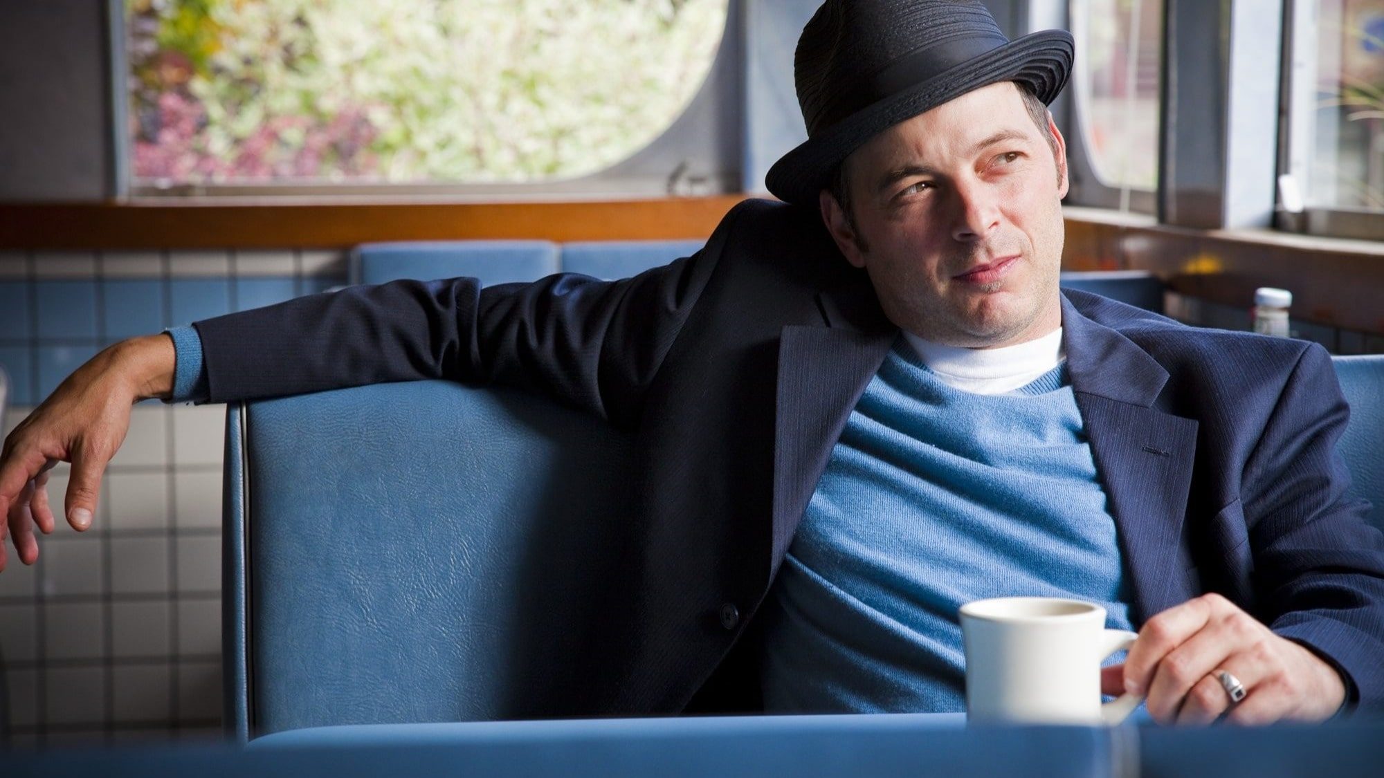 A man wearing a black fedora and a suit jacket sits in a diner booth, holding a white mug. He looks out of the window with a thoughtful expression, as if wondering if something is going on outside.