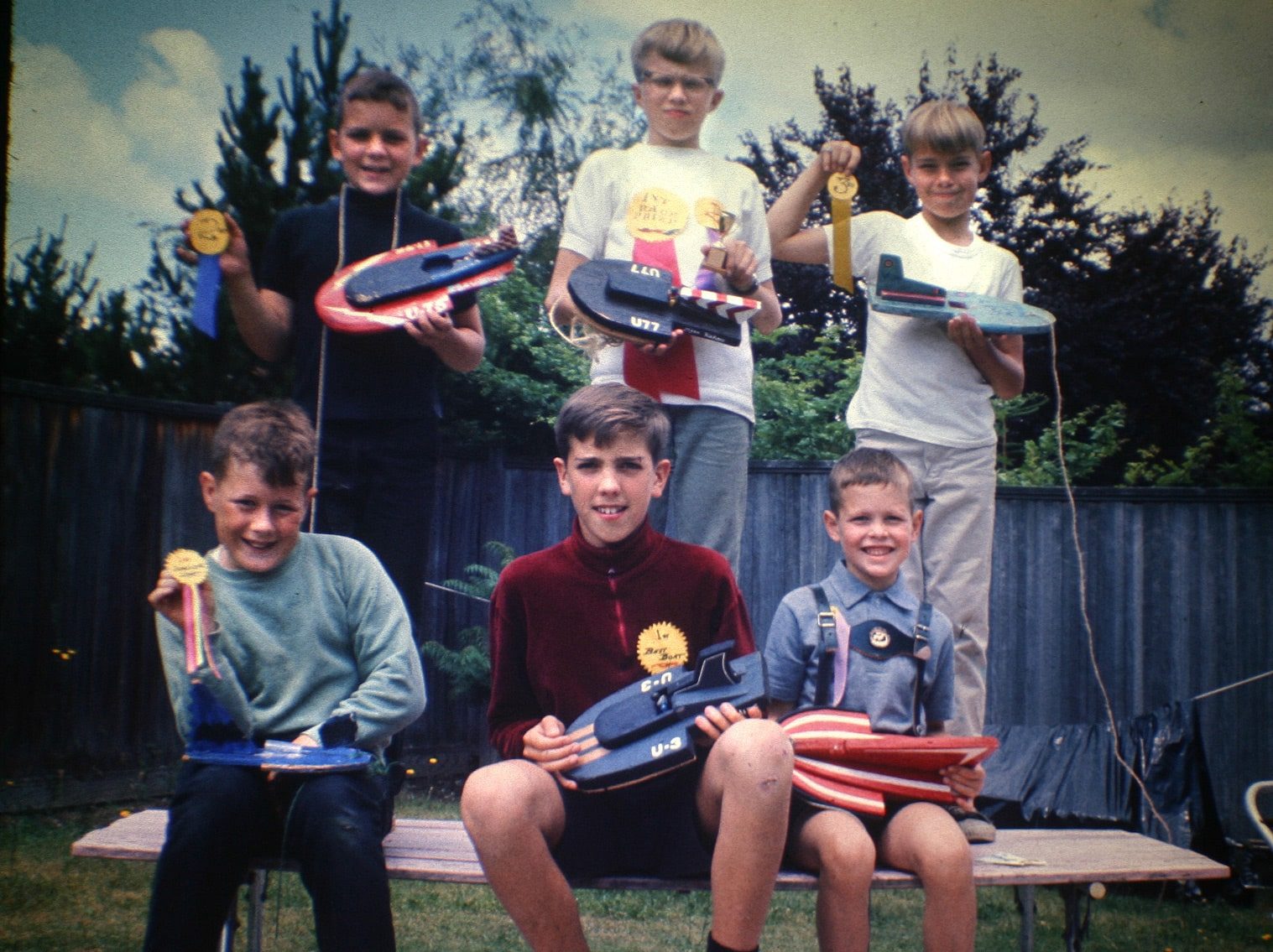 Five boys proudly display toy hydroplanes and wear medals, standing and sitting on a bench in a fenced backyard. Trees and bushes frame the background.