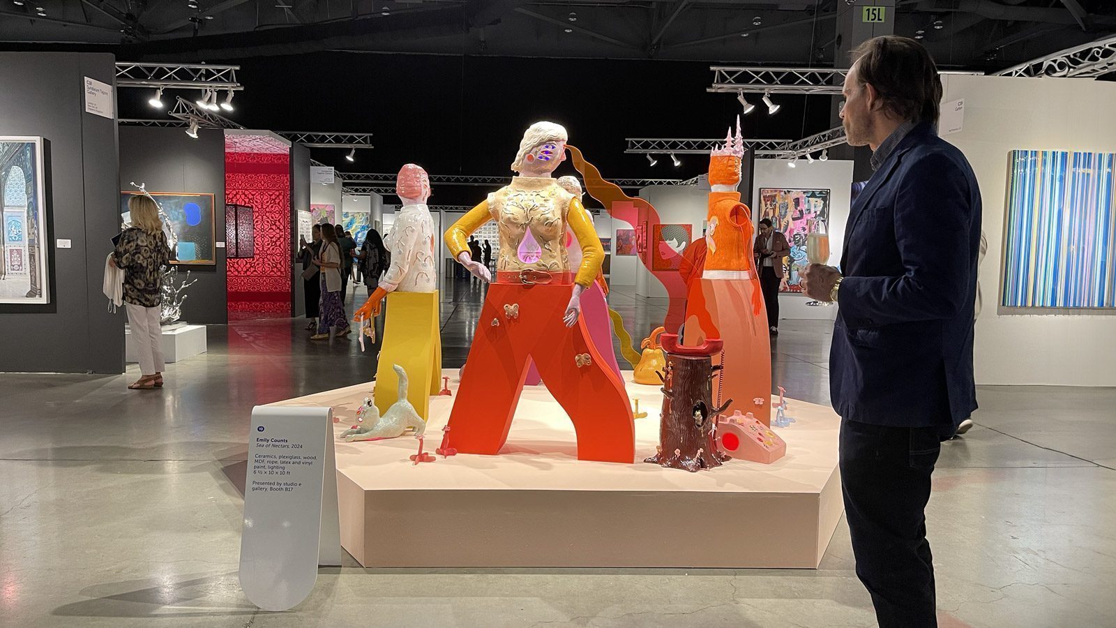Visitors observe colorful, abstract sculptures in a well-lit art gallery at the Seattle Art Fair. The sculptures are placed on a central platform with art pieces displayed on the surrounding walls, making for a strong showing of contemporary creativity.