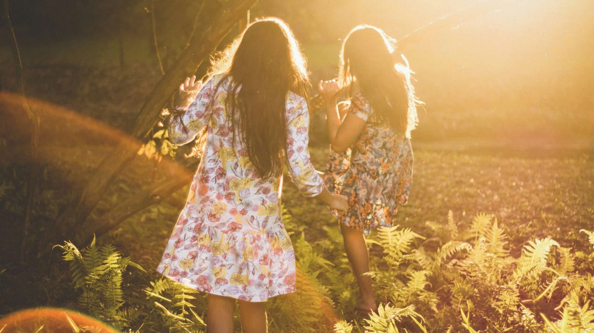 Two girls with long hair wearing floral dresses walk through a sunlit field with fern plants, radiating summer vibes.