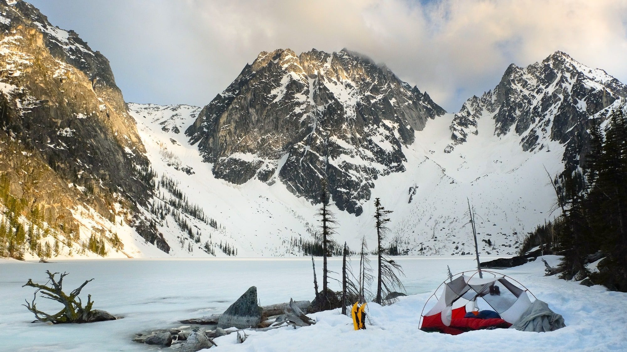 A red and gray tent is set up on snow near a frozen lake, surrounded by snow-covered mountains under a partly cloudy sky.