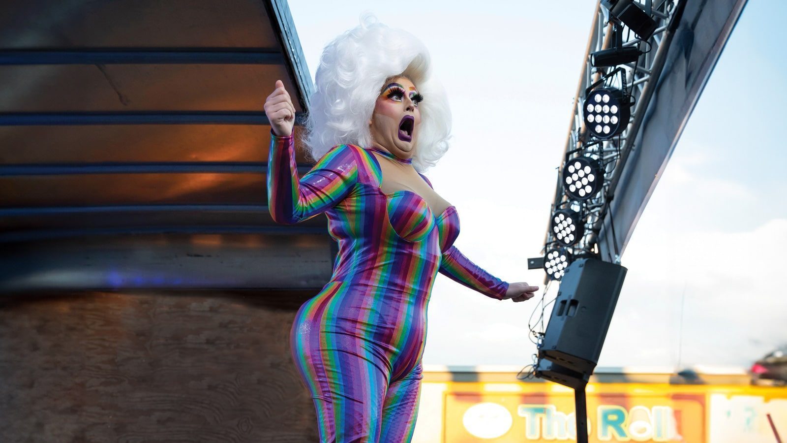 Person wearing a colorful striped outfit and a large, white wig performs on stage under studio lights.