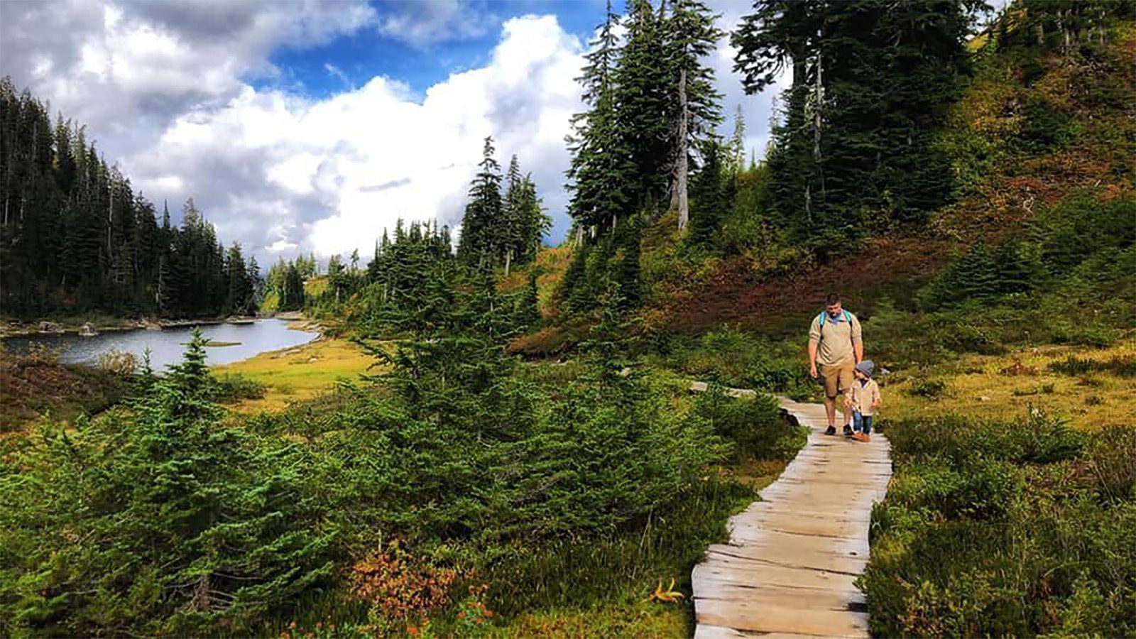 A person hikes with a child on a wooden path through a lush, green forested area, with a lake on the left side and a haze lingering under the cloudy sky above.