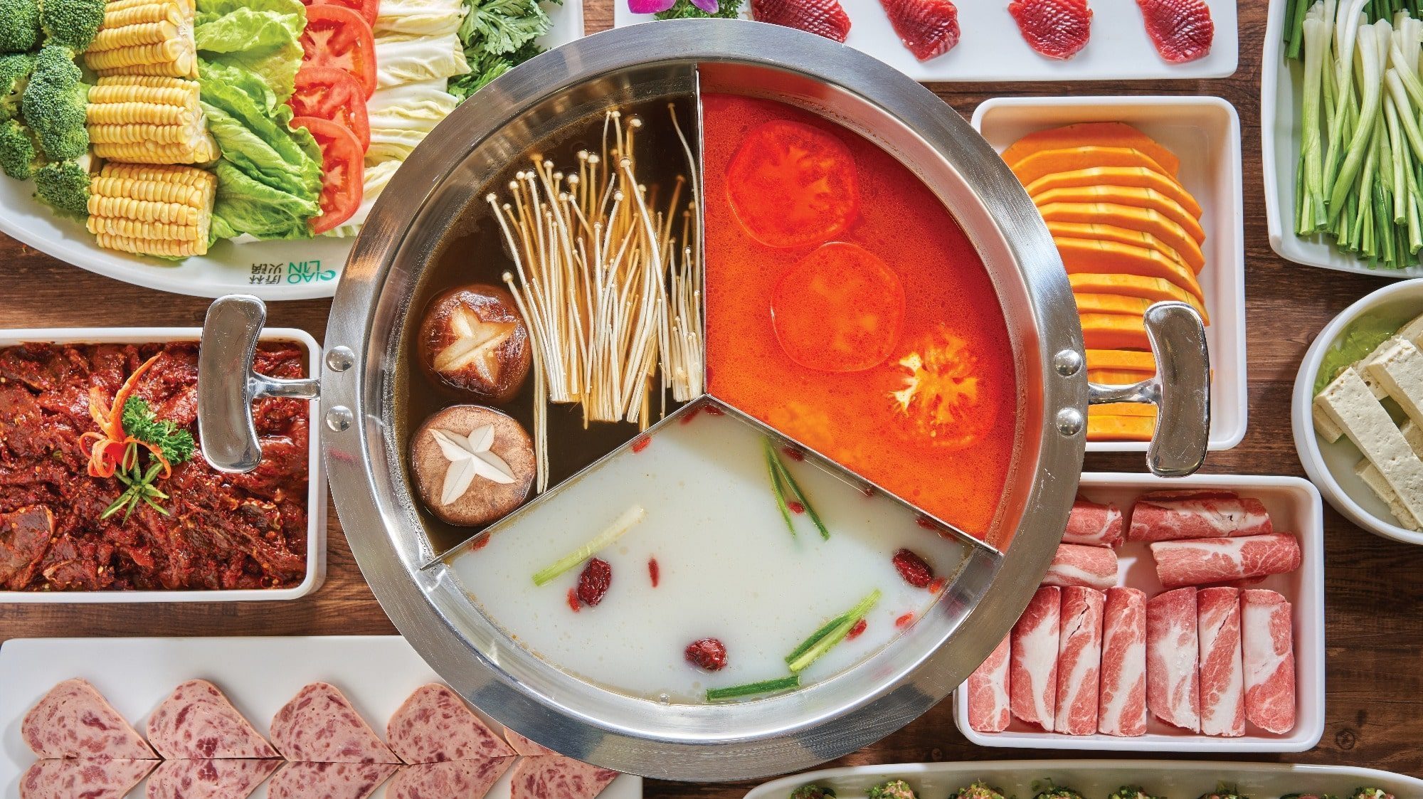 A four-section Sichuanese hot pot filled with different broths and ingredients, surrounded by plates of vegetables, meats, and side dishes.