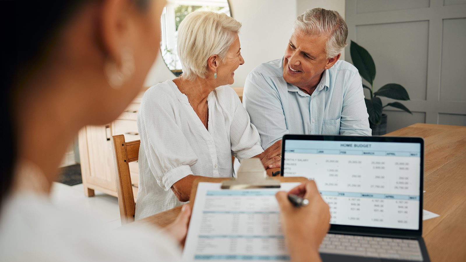 A senior couple sits and smiles at each other while a professional, showcasing a sense of style, presents them with a financial document and a laptop displaying their retiring home budget spreadsheet on the table.
