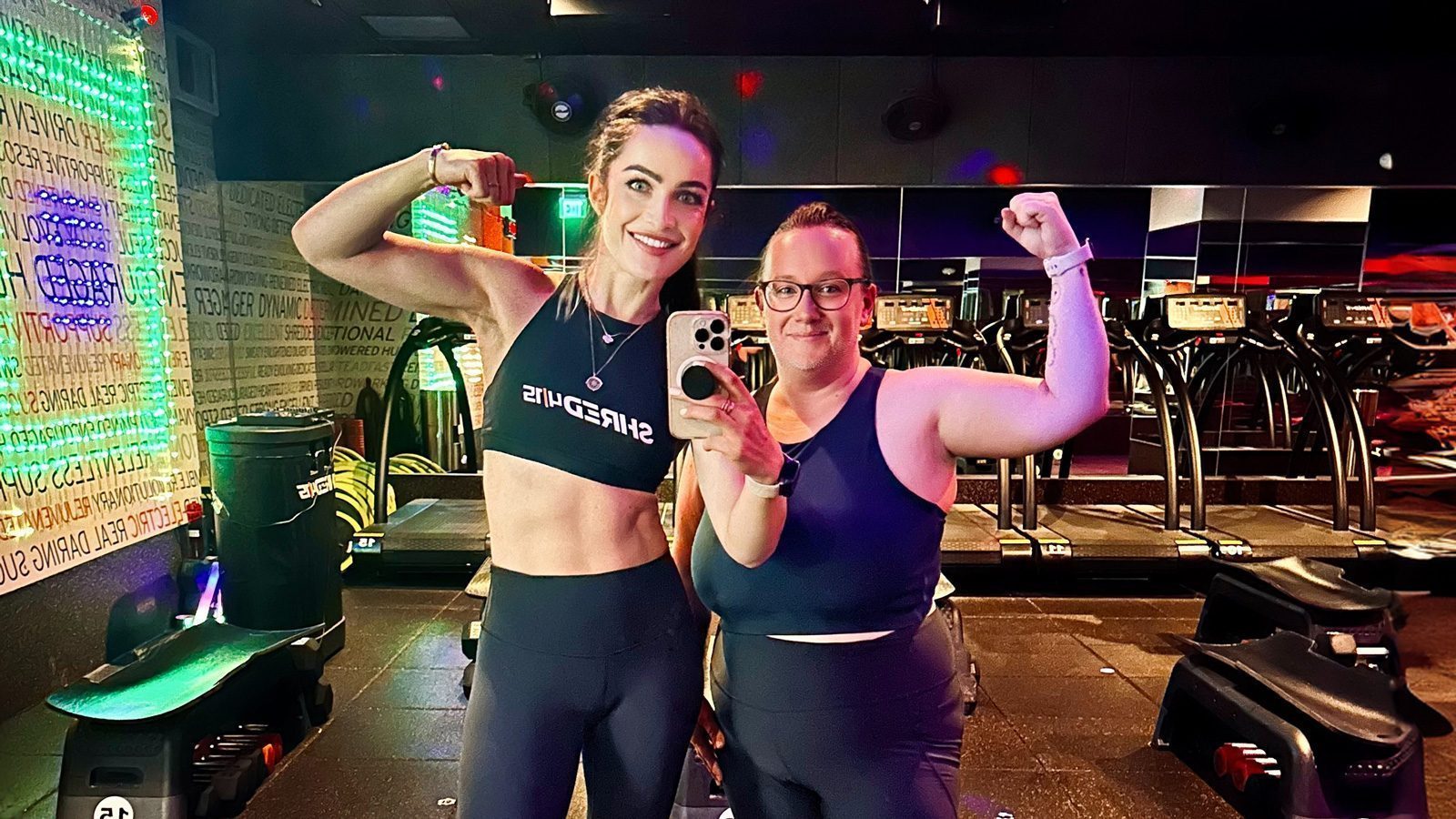 Two women pose in a gym, flexing their biceps and smiling at the camera, unlocking their fitness potential. One holds a phone, capturing the photo. Fitness equipment is visible in the background.