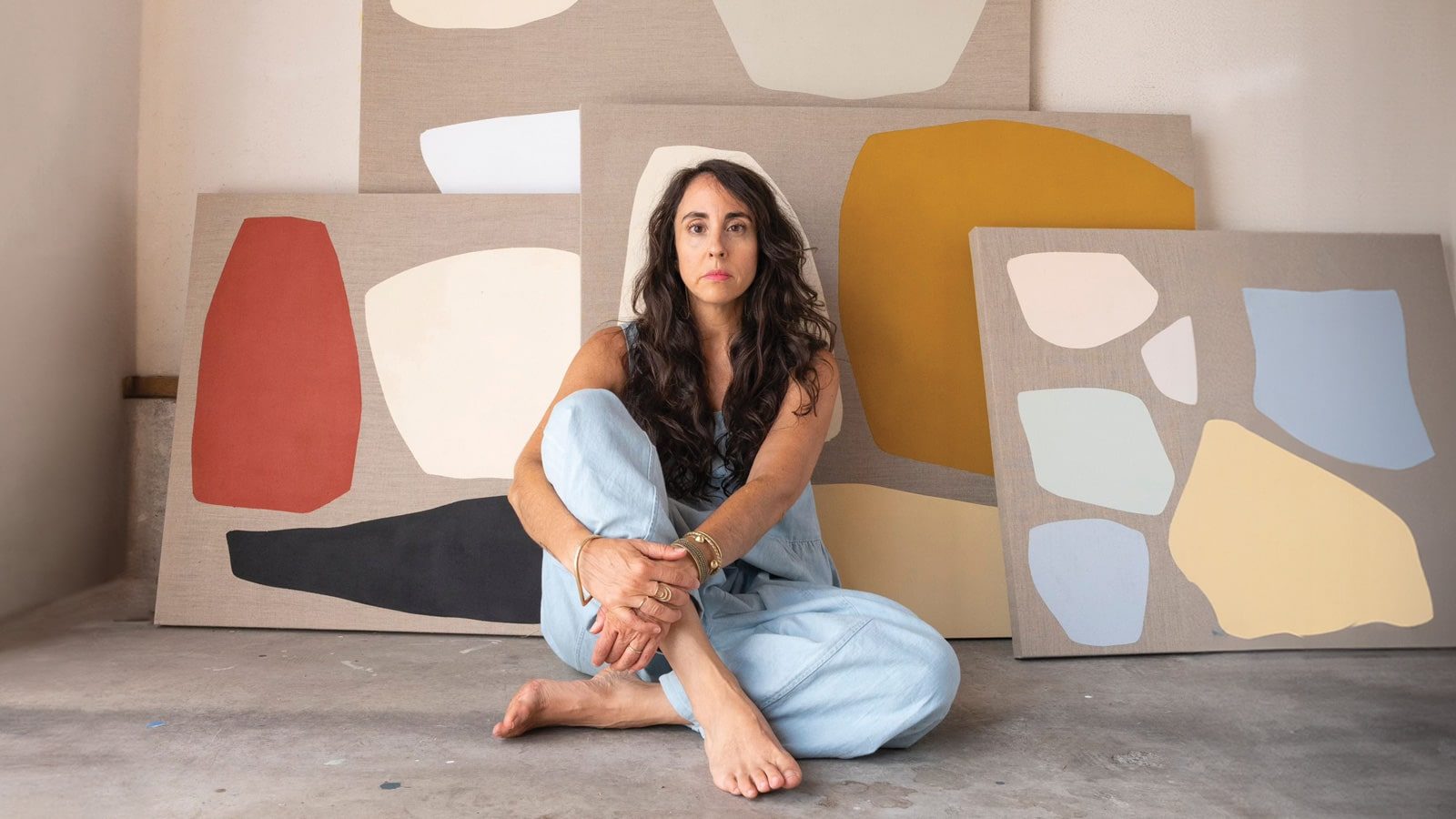 A woman sits on the floor in front of several abstract art pieces with geometric shapes in various colors. Embracing autumn hues, she has long hair and wears a light blue outfit, looking directly at the camera.