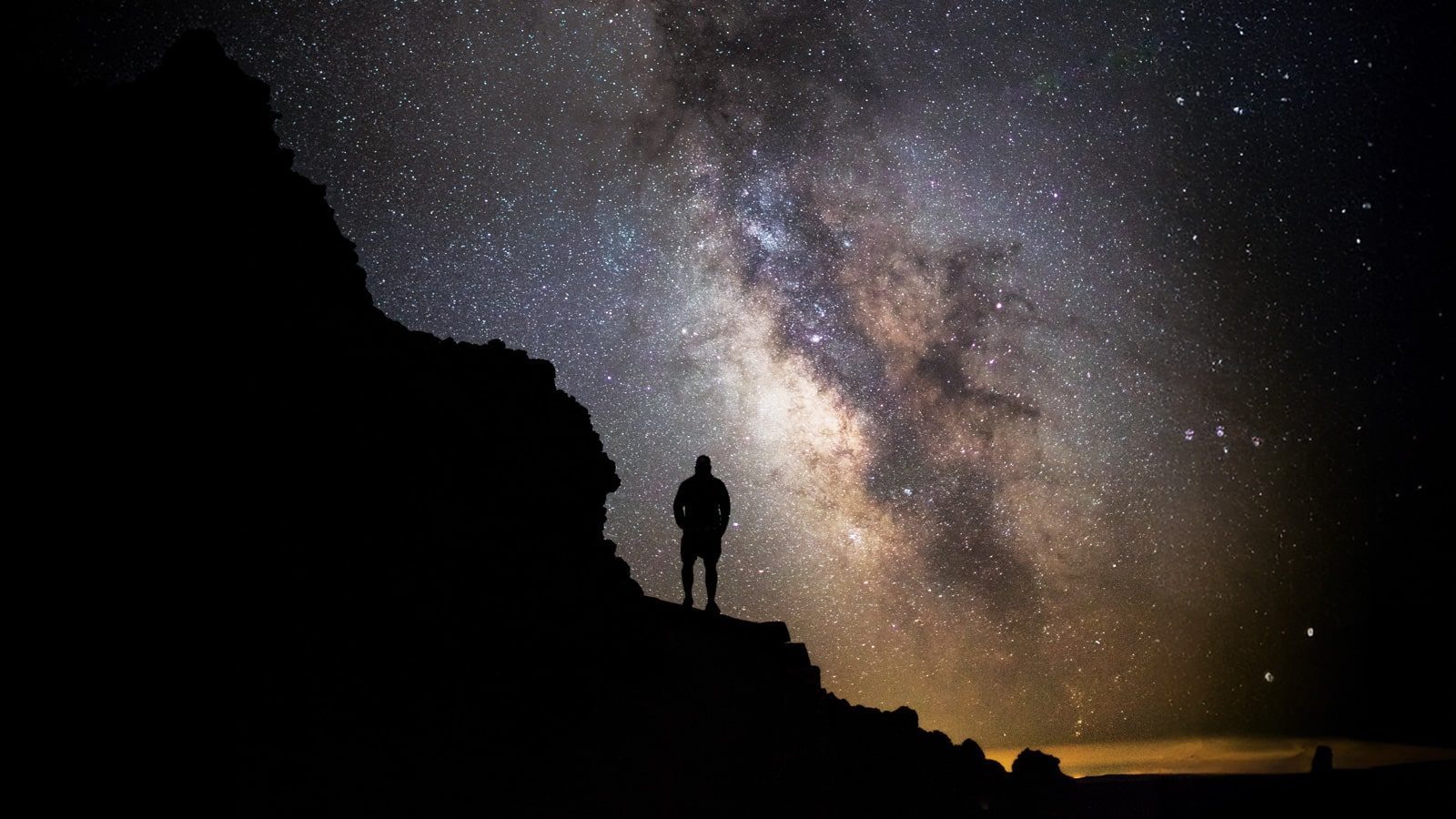 A silhouette of a person stands on a rocky terrain under a sky full of stars and the Milky Way galaxy, capturing the essence of autumn with its crisp, clear atmosphere—an essential moment for any stargazer.