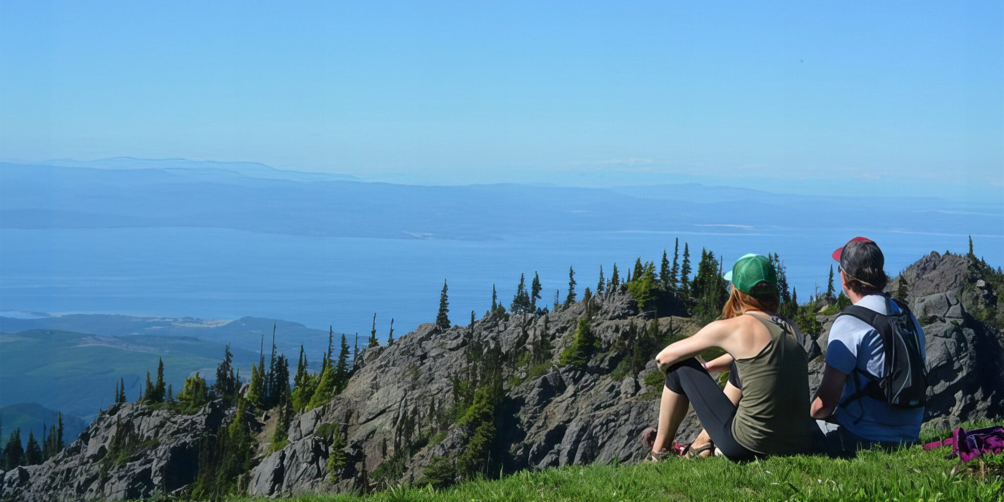 Two people sitting on a grassy area at the edge of a rocky hill, right outside overlooking a scenic view of mountains and a large body of water under a clear blue sky— truly one of nature's best kept secrets.