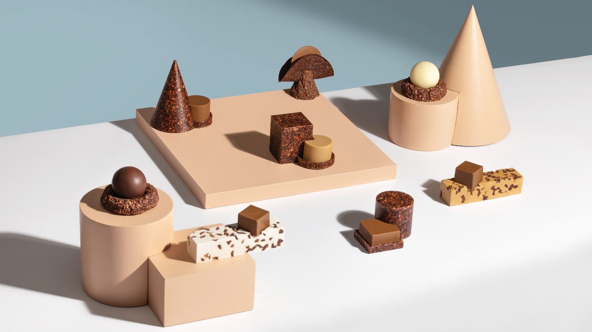 A geometrically arranged display of various chocolates and confections, featuring spheres, cones, cubes, and bars on beige and white platforms against a blue gradient background reminiscent of dark skies observed from distant observatories.