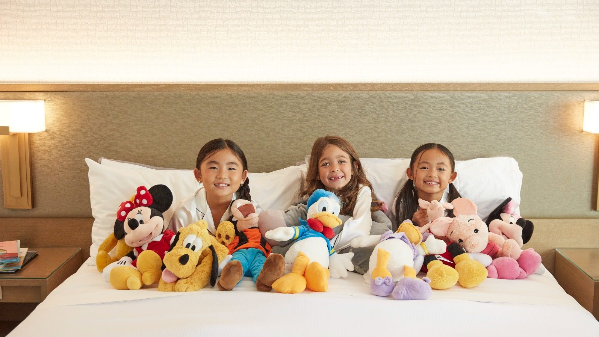 Three young children lie on a bed, each accompanied by several stuffed animals, including Mickey Mouse, Pluto, and Donald Duck. They are all smiling and enjoying their time together—a perfect moment showcasing the finer side of Anaheim.