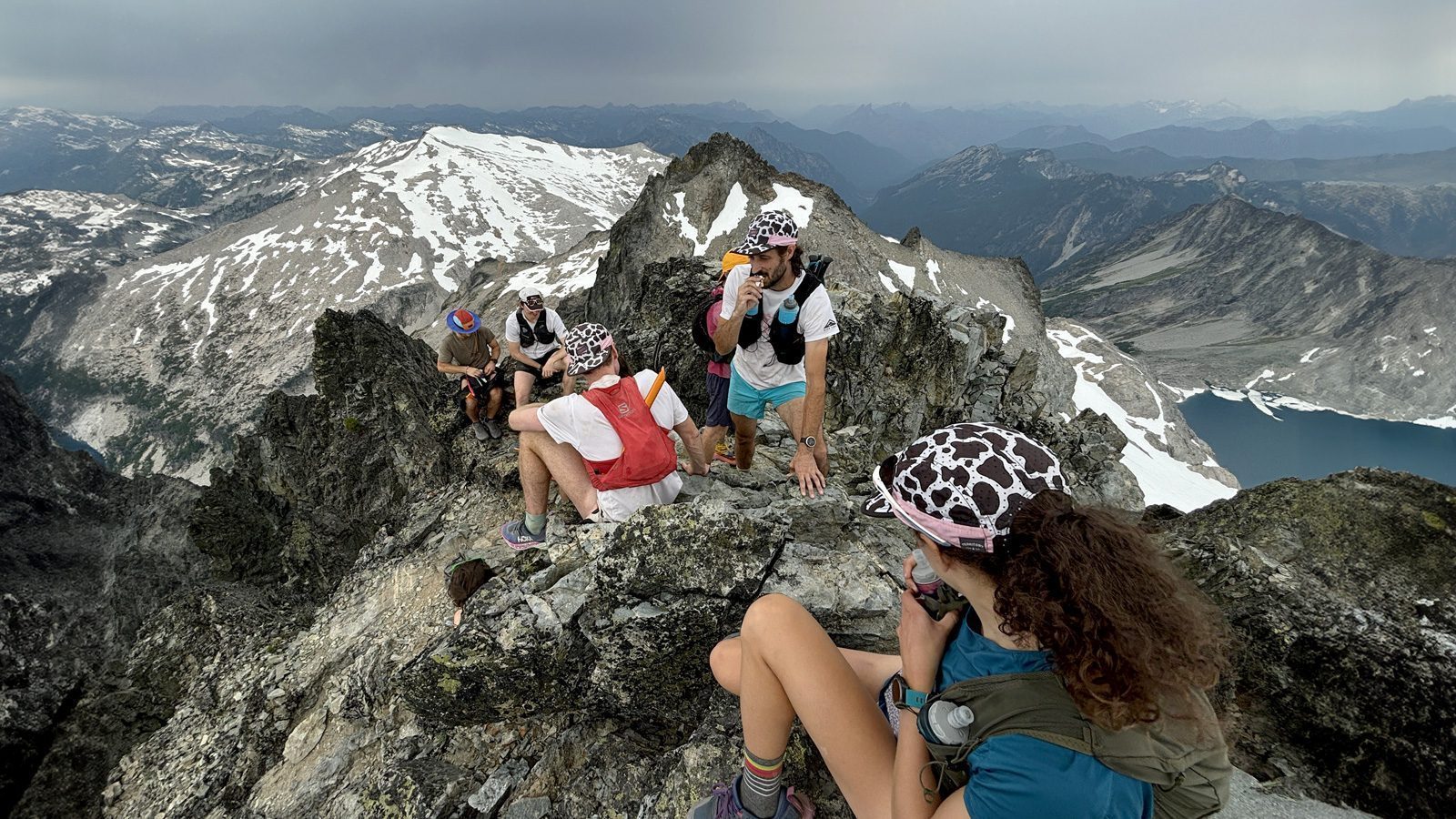 A group of hikers wearing similar caps rests on a rocky mountain peak with snow-covered slopes and distant mountains visible under a cloudy sky, savoring the off-road rush that brought them to this breathtaking vista.