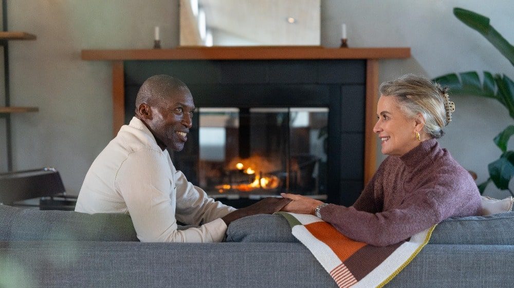 A man and woman sit on a couch facing each other, smiling and holding hands in front of a fireplace in a cozy living room, nestled in the heart of Oregon's Tualatin Valley.