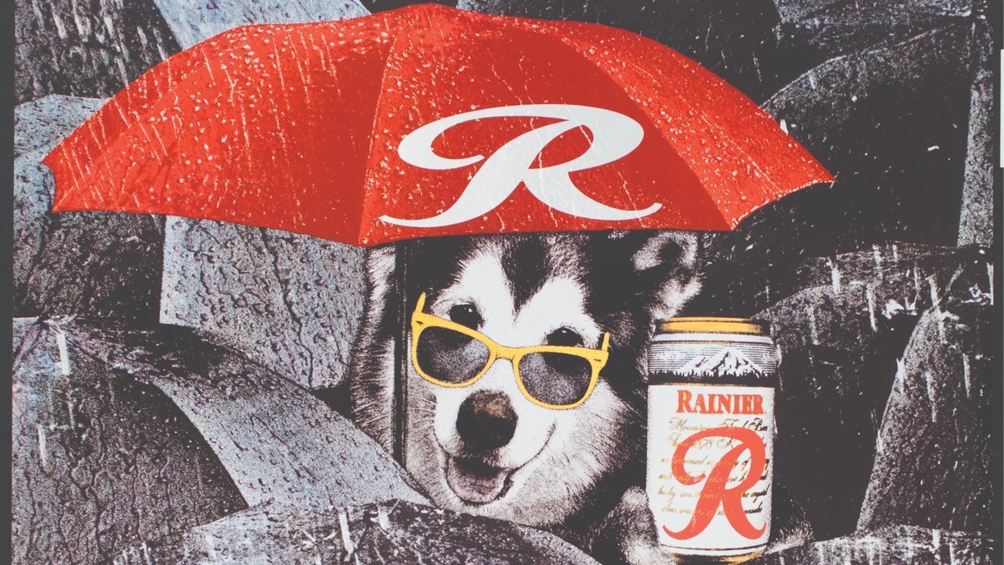 A dog wearing yellow sunglasses sits under a red umbrella, holding a can of Rainier beer against a rocky background. The crisp can of Rainier beer glistens in the sunlight, adding to the scene's charm.