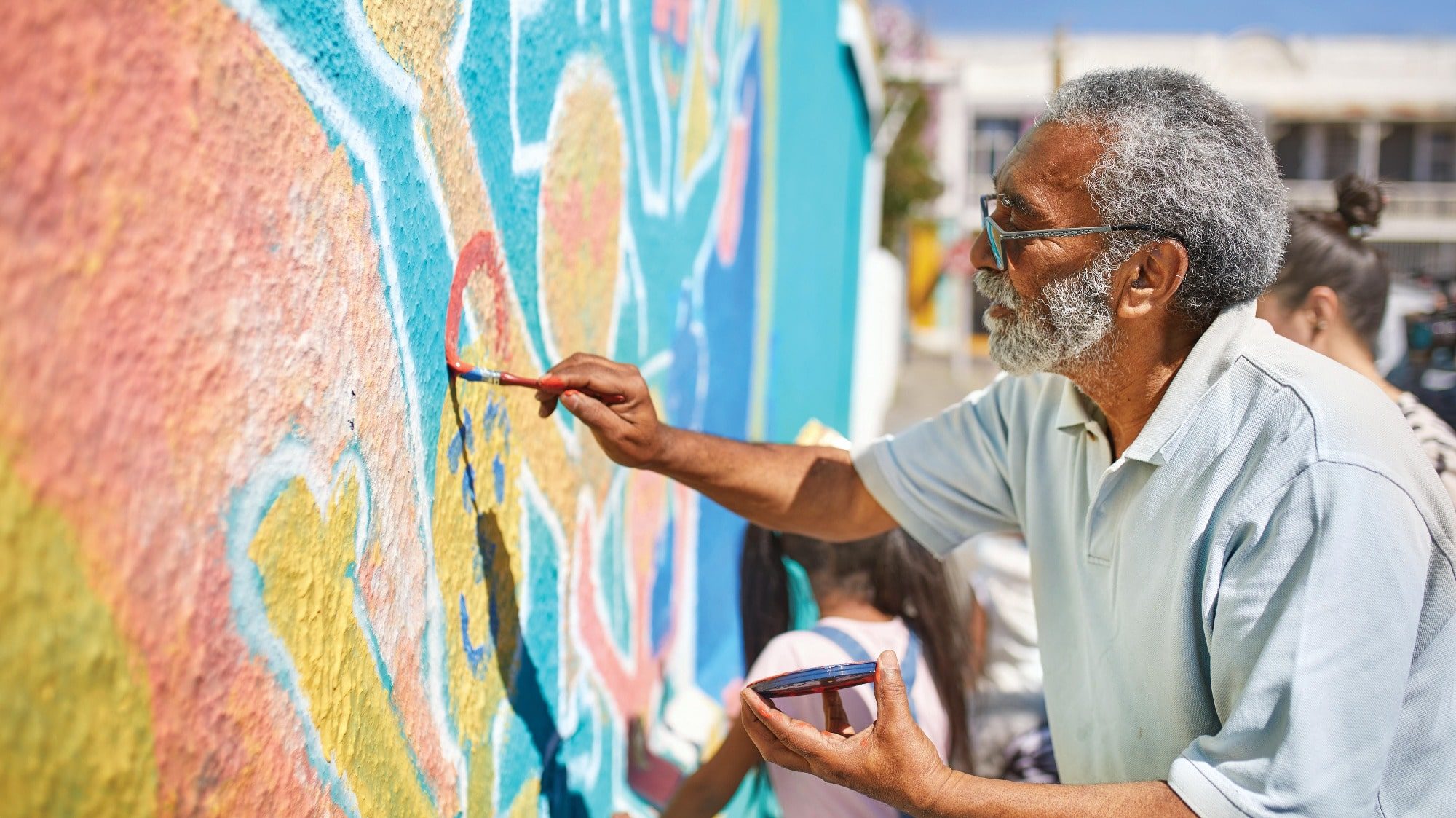 An elderly man paints a colorful mural on a wall outdoors in the Land of Enchantment. He is holding a paintbrush and a palette, while a child works beside him. This vibrant scene embodies the artistic spirit of New Mexico, creating lasting travel memories for those who witness it.