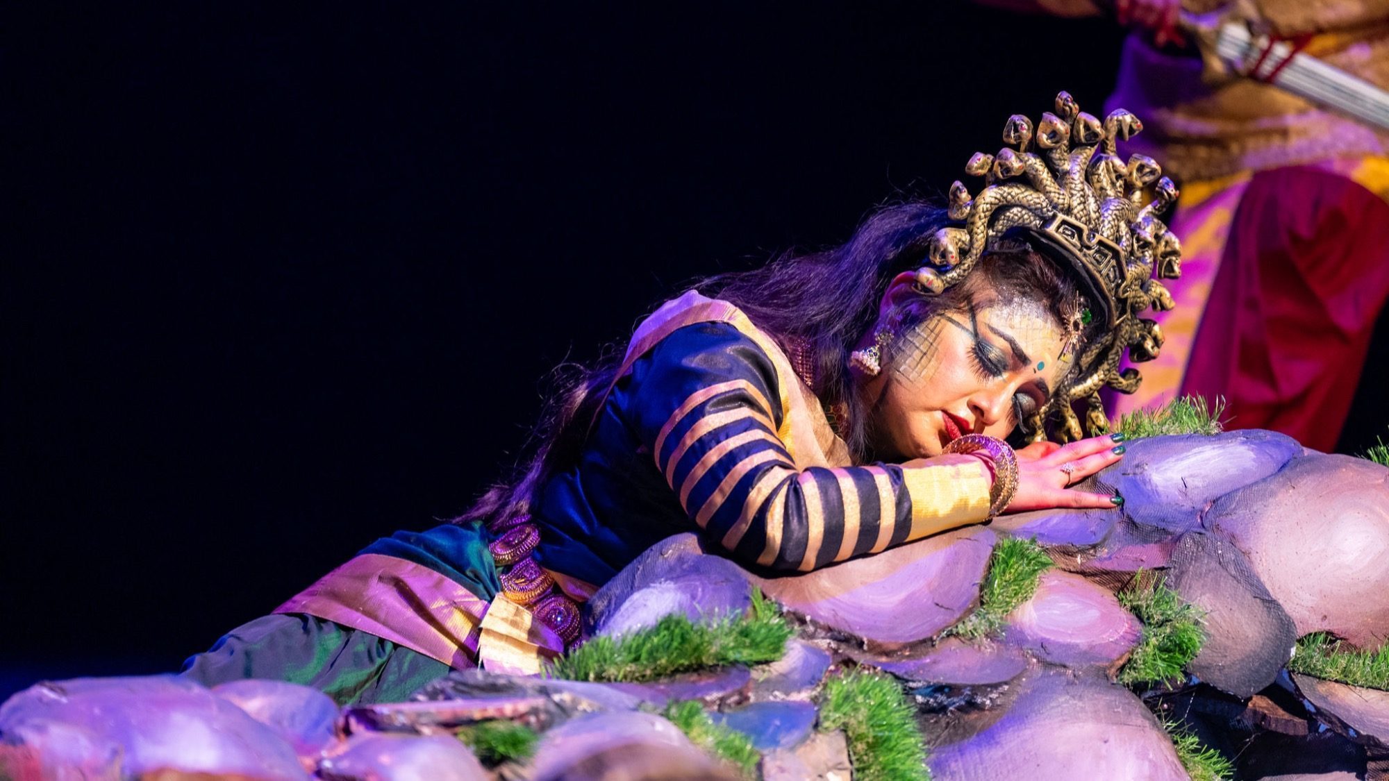 A person in elaborate costume and makeup, wearing a headdress, lies on decorative rocks during a stage performance of Cleopatra, appearing to be asleep.