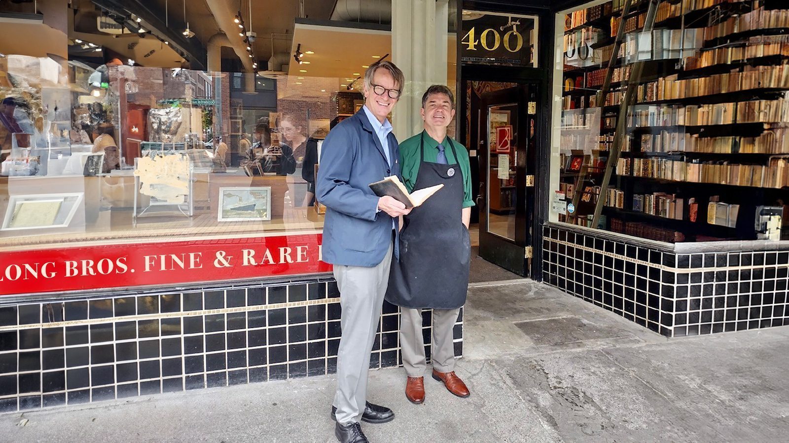 Two people stand outside a bookstore. One holds a book, and the other wears an apron. Shelves of books are visible inside, and a "Fine & Rare" sign is displayed on the window.