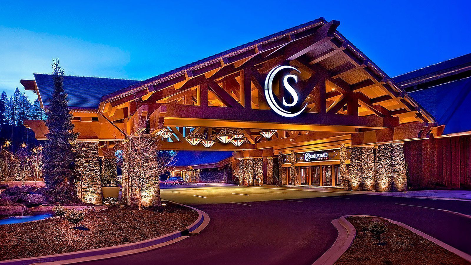 Exterior view of a building with a large entrance, featuring a wooden structure and a circular sign with the letter 'S'. The Snoqualmie Casino is beautifully illuminated at night, adding to its allure as possibly the best casino experience.