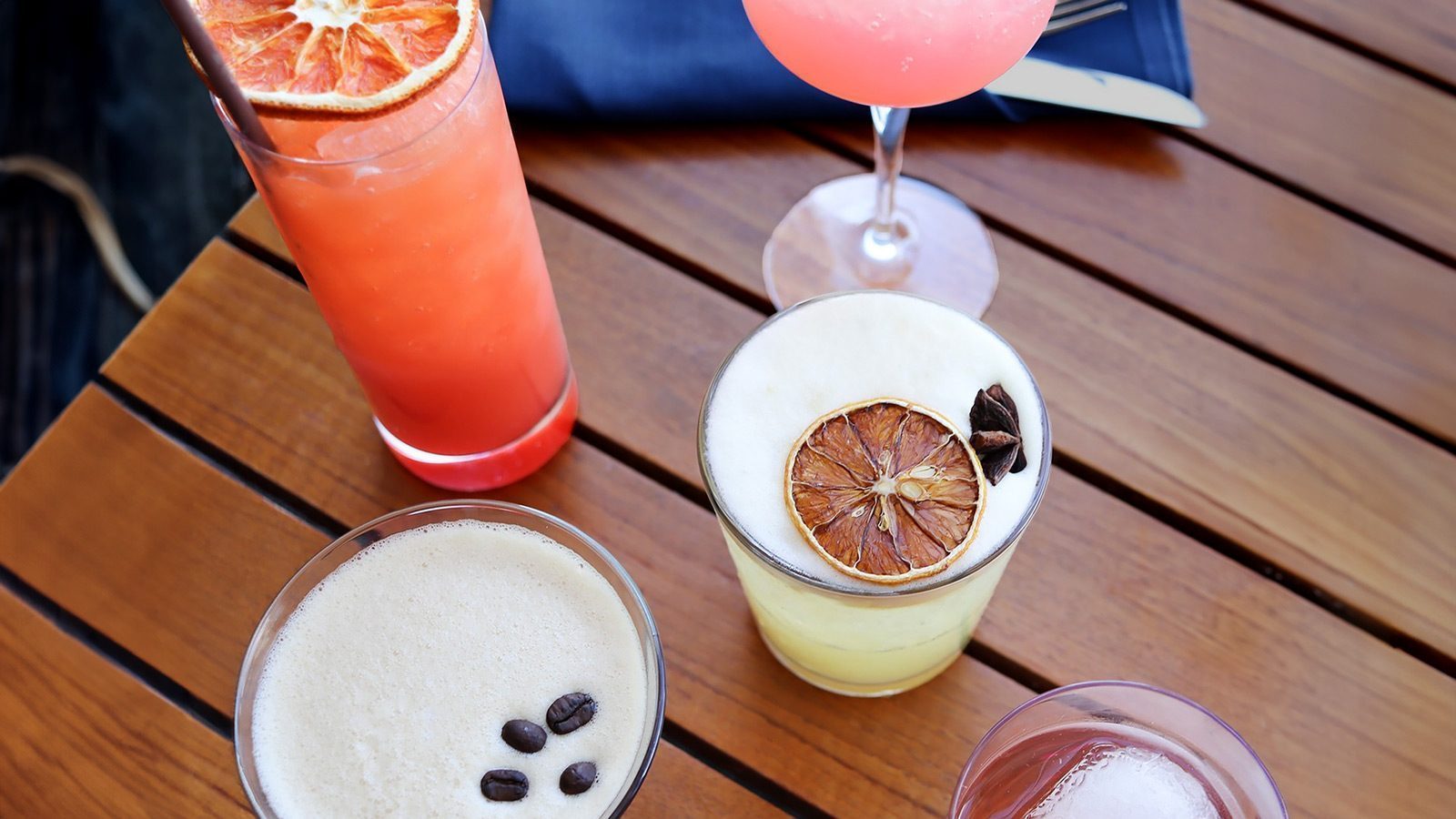 An overhead view of assorted craft cocktails on a wooden table, featuring drinks with garnishes like dried citrus slices, coffee beans, and a cloth napkin with cutlery in the background.