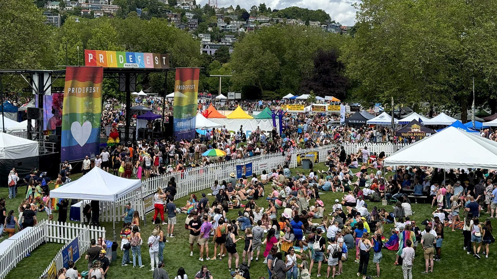 A vibrant scene at Seattle PrideFest, celebrated as the best festival, shows crowds gathered around colorful tents and two main stages with rainbow banners, set in a park surrounded by trees and distant buildings.