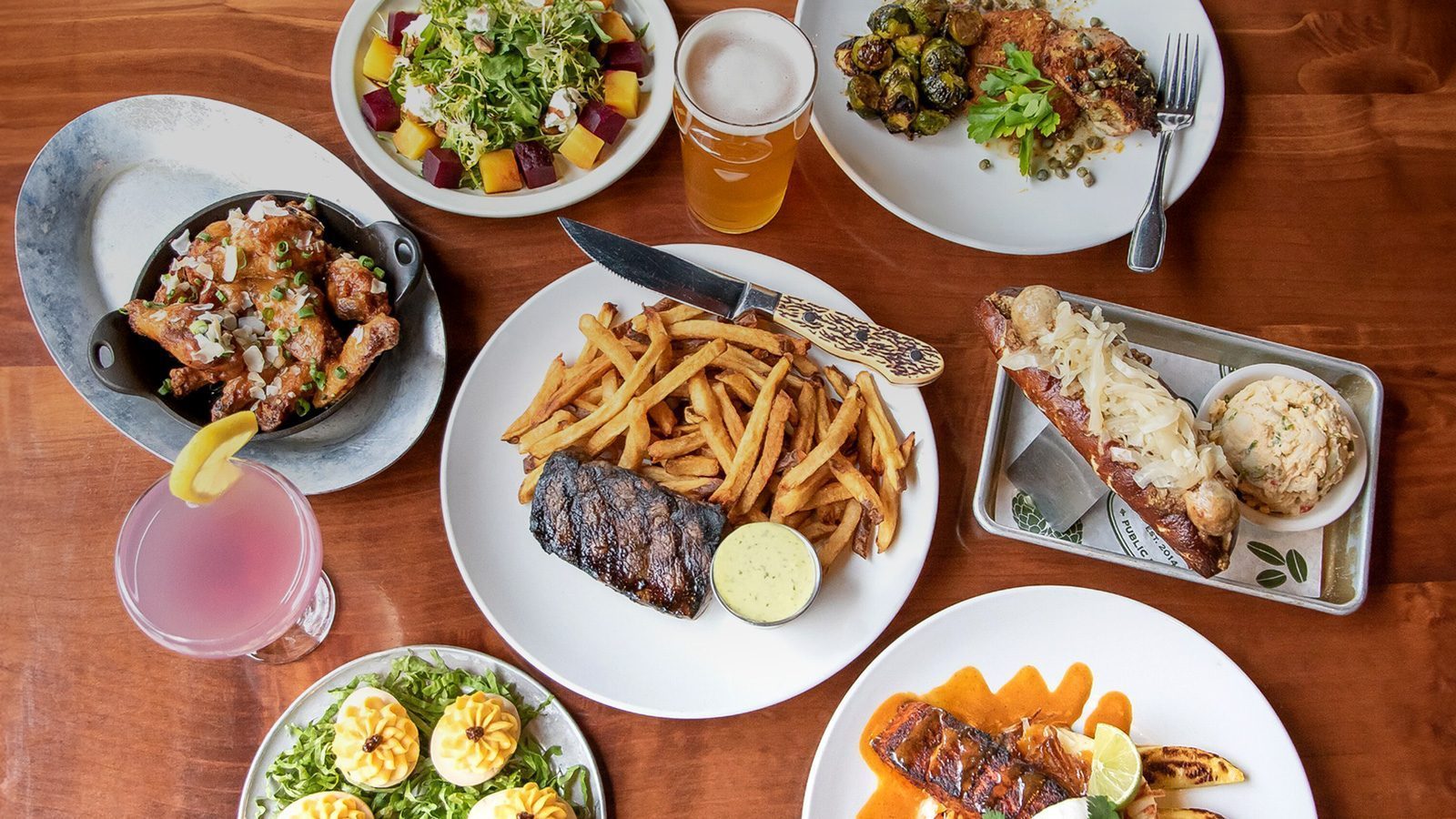 At our neighborhood restaurant, Beardslee Public House in Bothell, the table is a feast of delights: succulent steak and fries, deviled eggs, mouthwatering hot dogs, crispy chicken wings, fresh salad, roasted vegetables, cold beer, and a vibrant pink cocktail. Experience the best culinary offerings around!