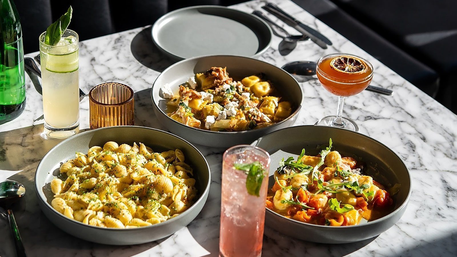 Three bowls of pasta sit on a marble table at the new restaurant, accompanied by two cocktails and a glass of water, with utensils nearby.