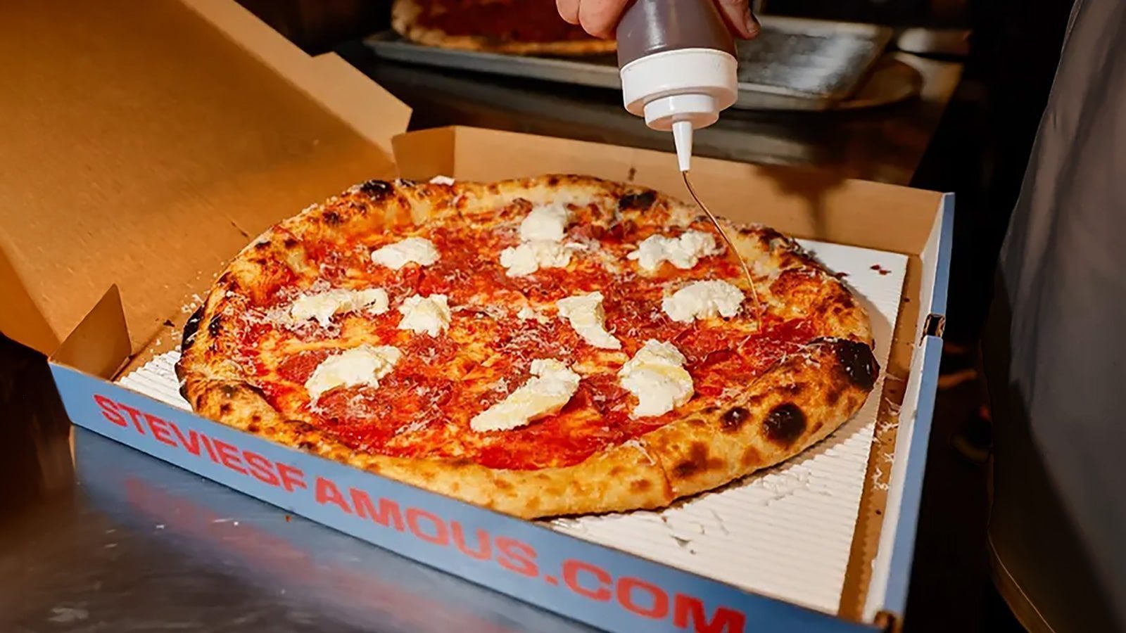 A pizza in a delivery box is topped with dollops of white cheese, while a sauce is expertly drizzled over it from a bottle—a signature touch from one of the exciting new Seattle restaurants.