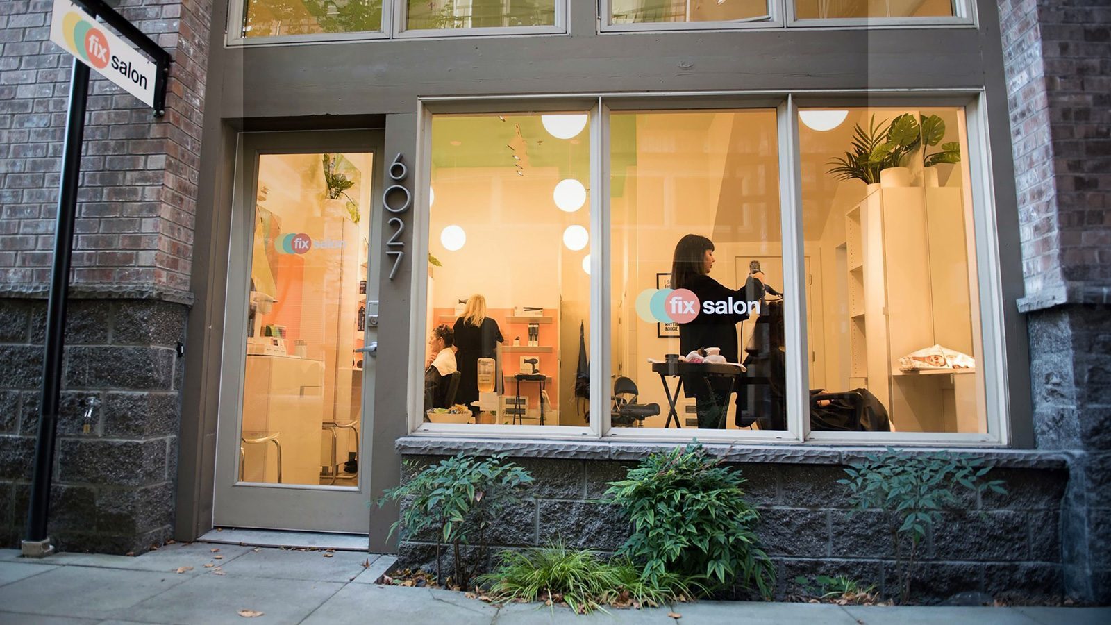 A vibrant street view showcases the bustling Fix Salon, where people can be seen through its large windows. The entrance is adorned with lush plants, inviting passersby into the chic oasis.