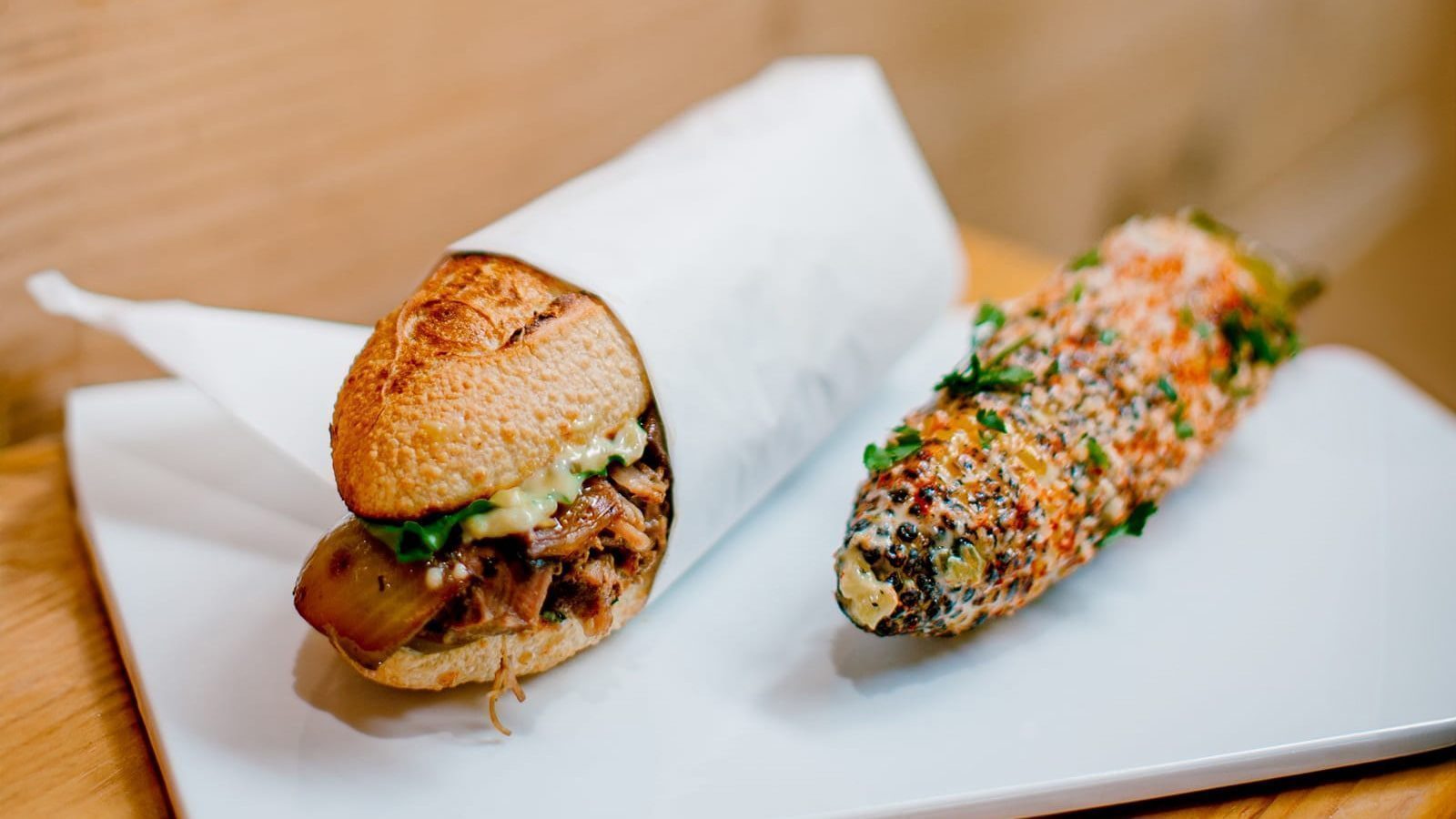 A sandwich with meat and sauce sits next to a seasoned corn on the cob, both elegantly presented on a white plate at Seattle's new restaurant delighting food enthusiasts.
