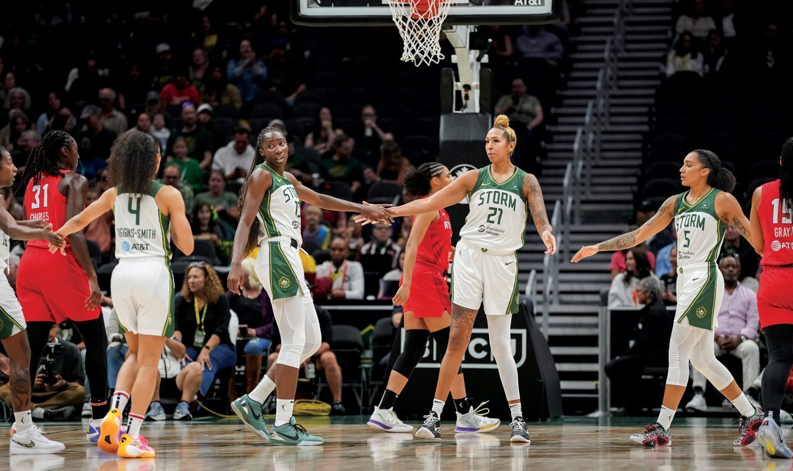 Players clad in "Storm" uniforms exchange high-fives on the basketball court as members of the opposing sports team, in red attire, look on. The stadium seats are partly filled with spectators, rooting for their favorite teams, including the spirited Seattle Storm.