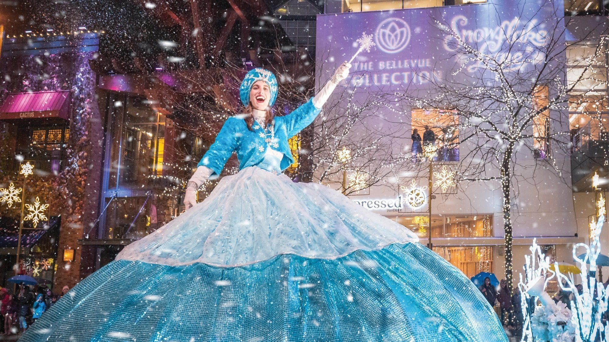 A performer in a blue dress dances outdoors in a snowy setting adorned with festive decorations and a sign reading "Snowflake Lane," showcasing what to do in Seattle during the holidays.
